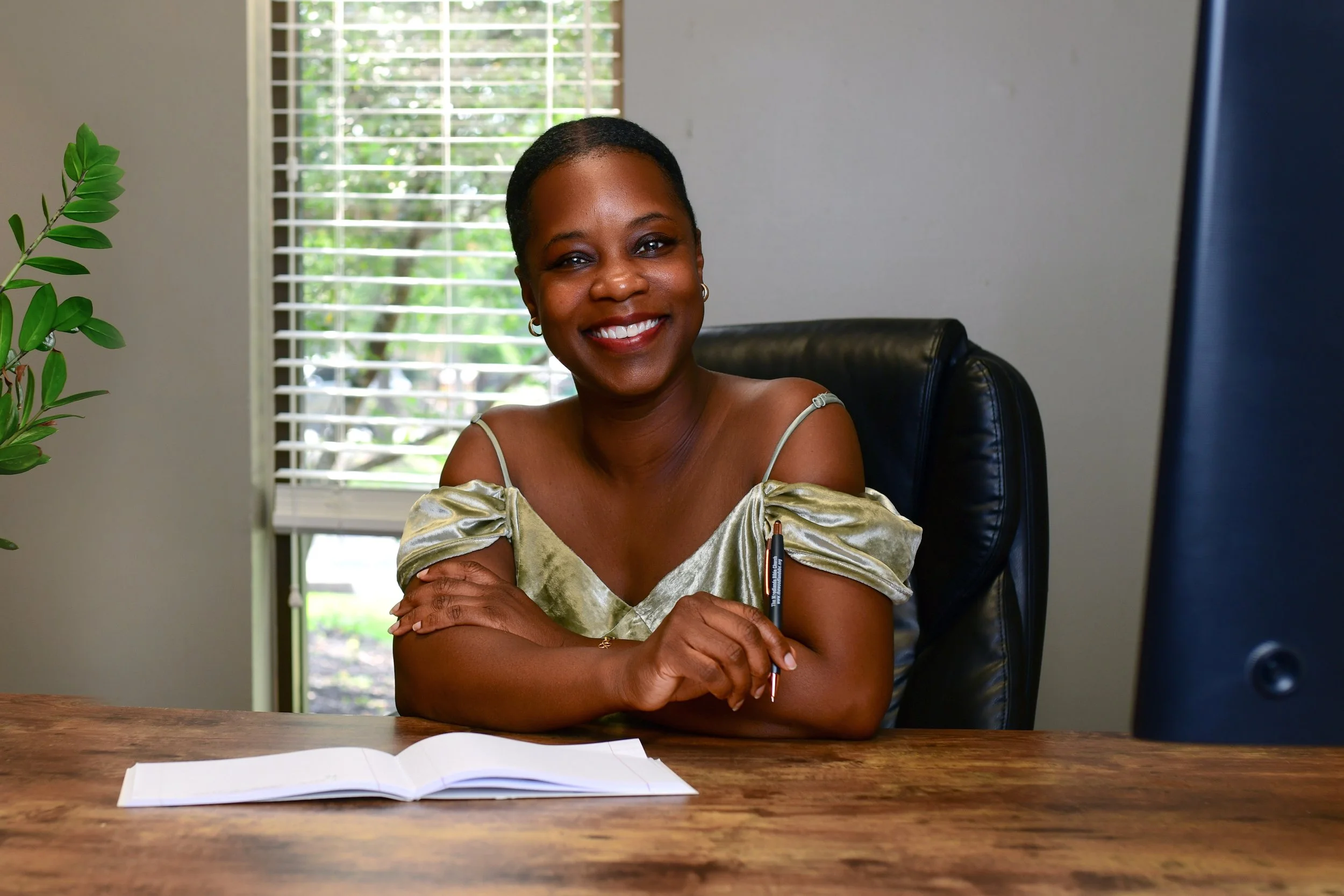 A woman sitting at a wooden desk, smiling while holding a pen, with an open notebook in front of her and a window with white blinds behind her.