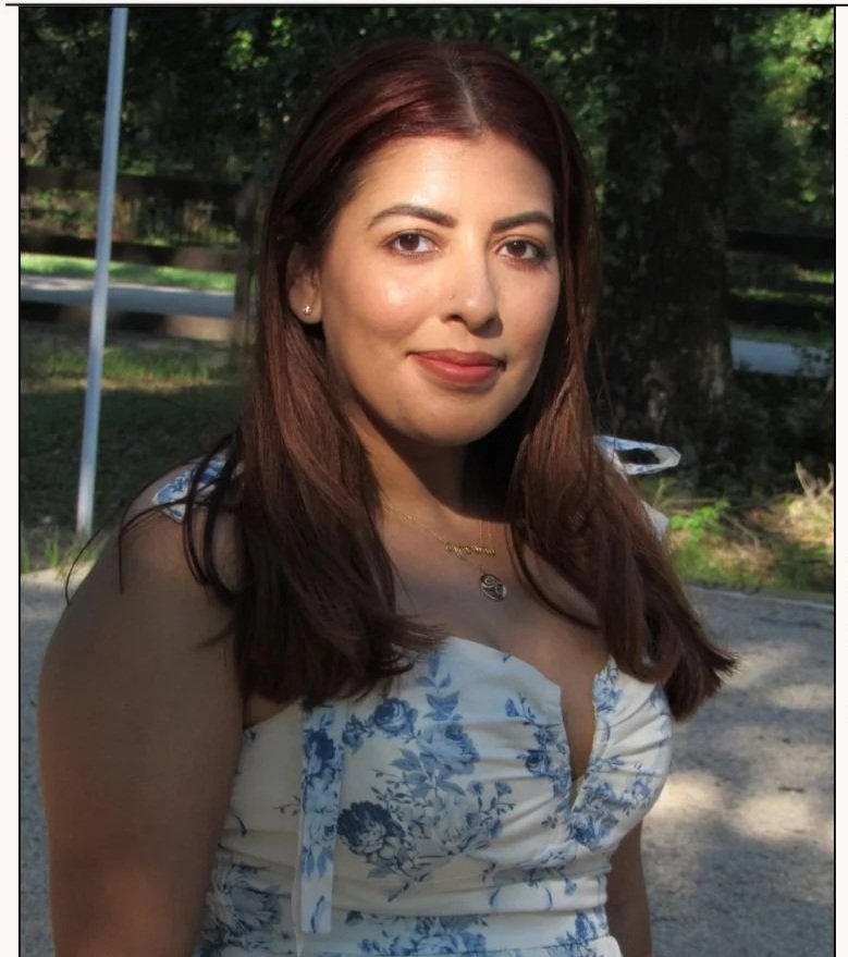 A woman with long auburn hair poses outdoors in daylight, wearing a white dress with blue floral patterns and a necklace, with trees and greenery in the background.