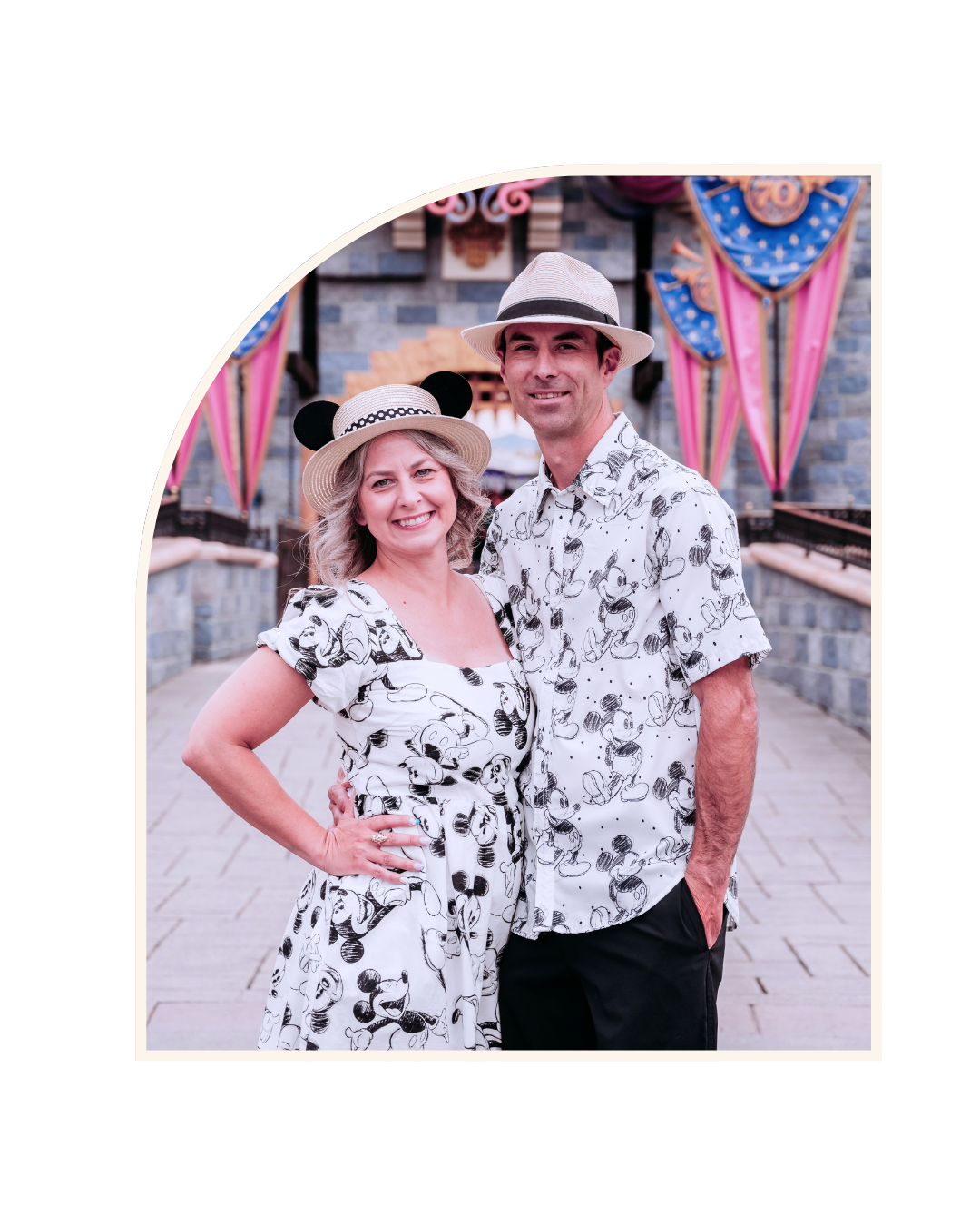 A smiling woman and man standing close together at a Disney-themed event, wearing matching Mickey Mouse print clothing and hats, with colorful banners and castle stone walls in the background.