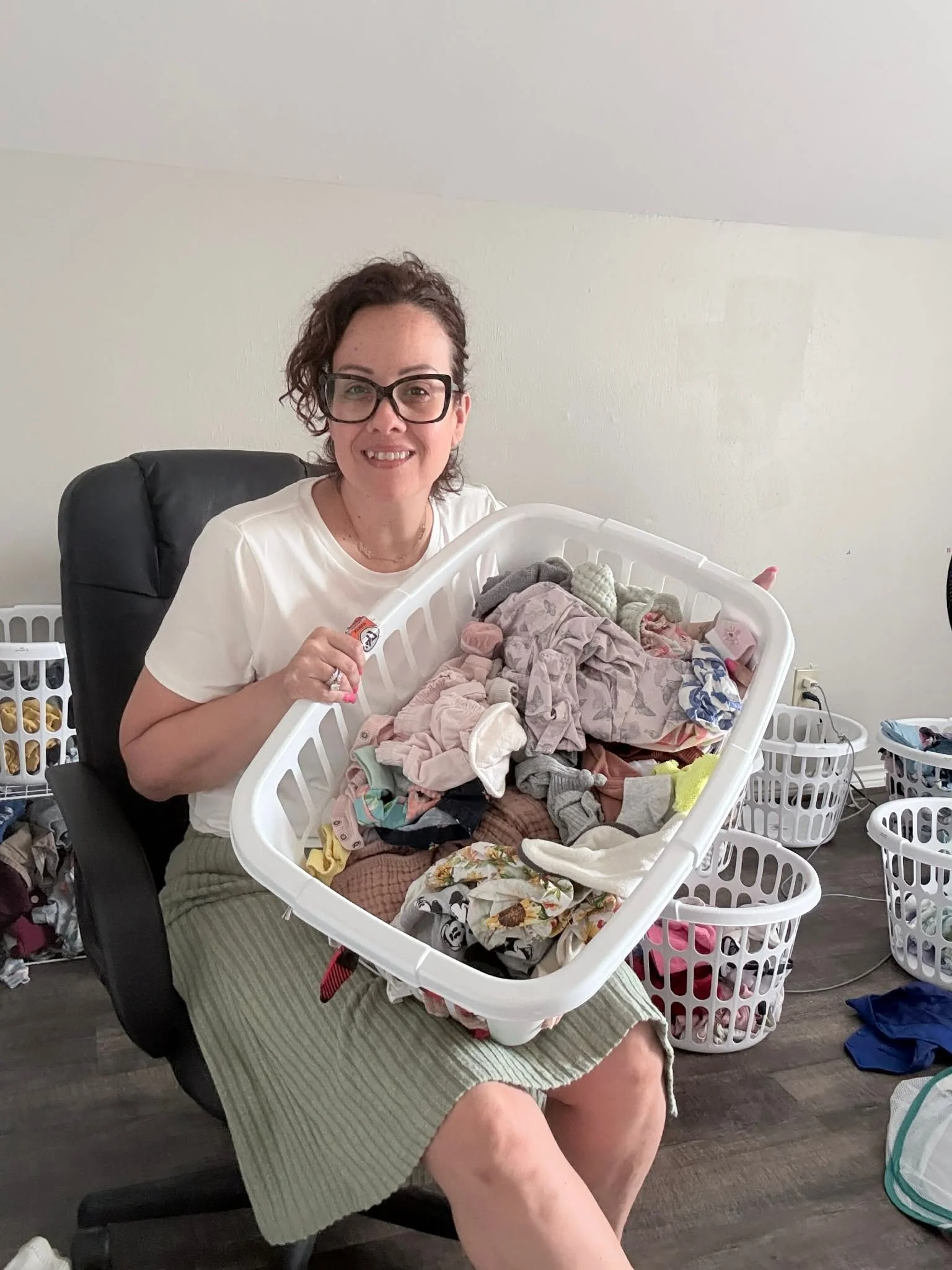 A woman wearing glasses and a white T-shirt, sitting on a chair, holding a laundry basket filled with various clothes, surrounded by laundry baskets and clothes on the floor in a room with a gray carpet and white walls.