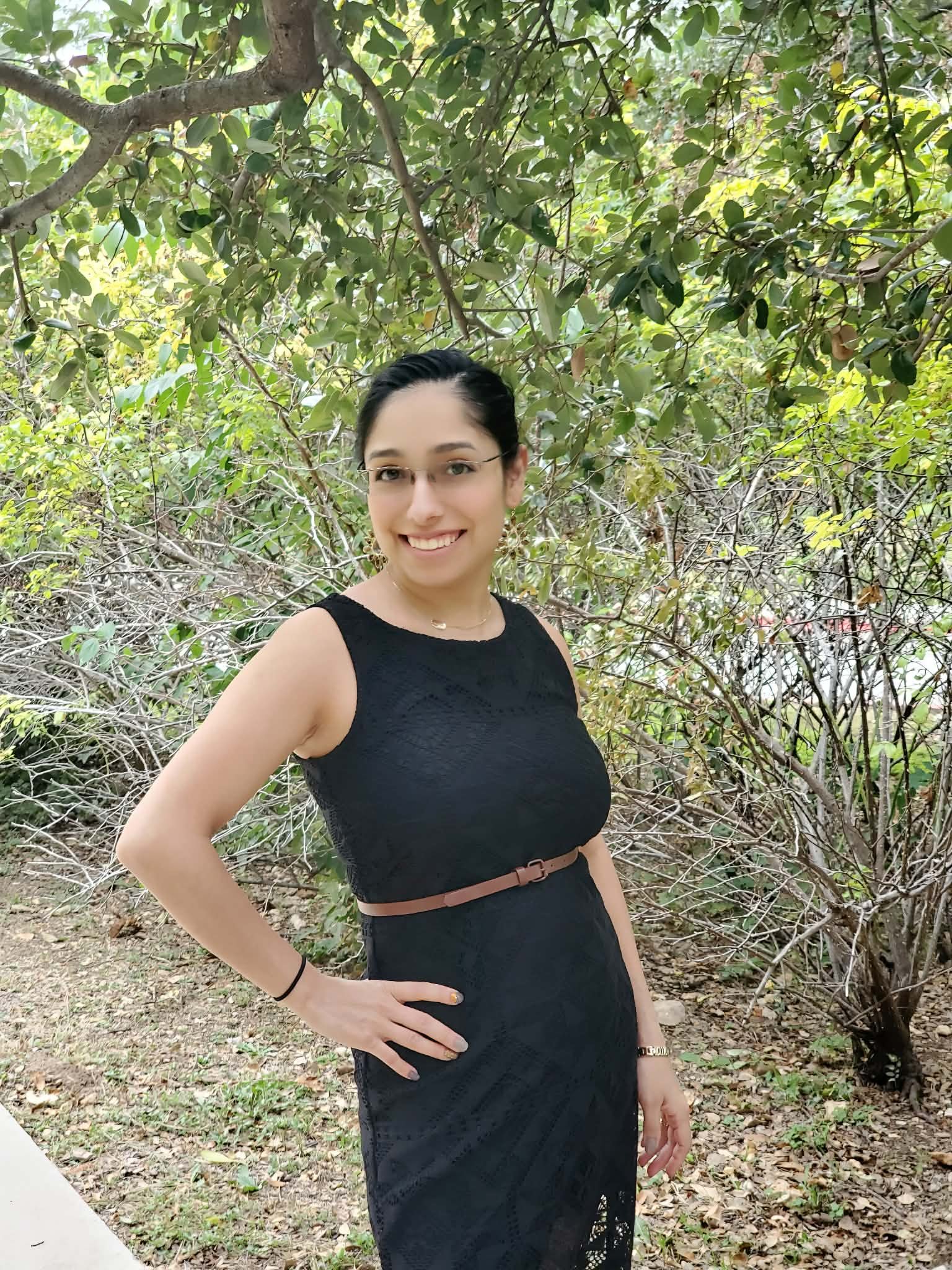 A smiling woman with glasses and dark hair tied back, wearing a black sleeveless dress and jewelry, standing outdoors with trees and leaves in the background.
