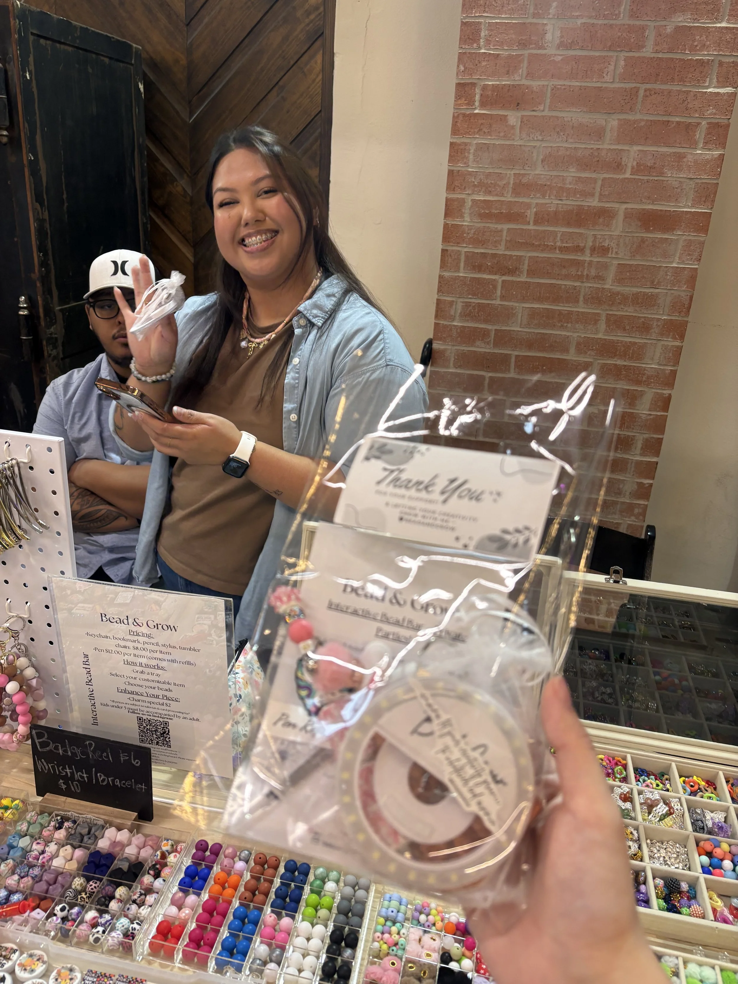 A woman with long dark hair, smiling, holding a small package of beads at a craft booth. Behind her is a man with glasses, wearing a white cap and gray shirt, looking in her direction. The booth has various beads in containers and a sign with prices 