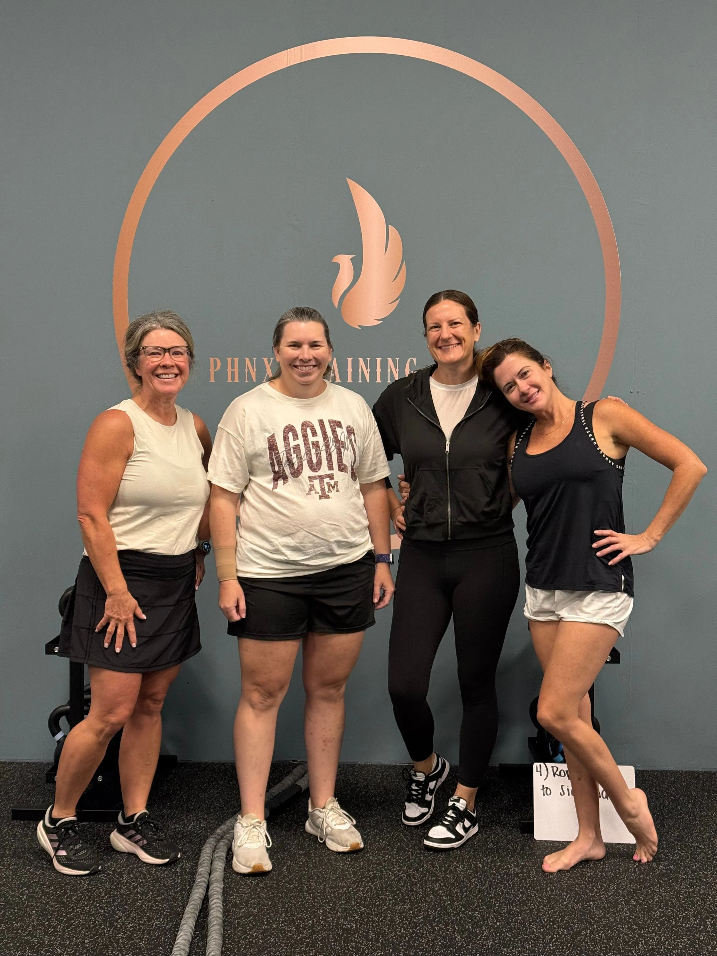 Four women standing together in a gym, smiling at the camera, with a wall logo behind them that reads 'PHOENIX TRAINING.'