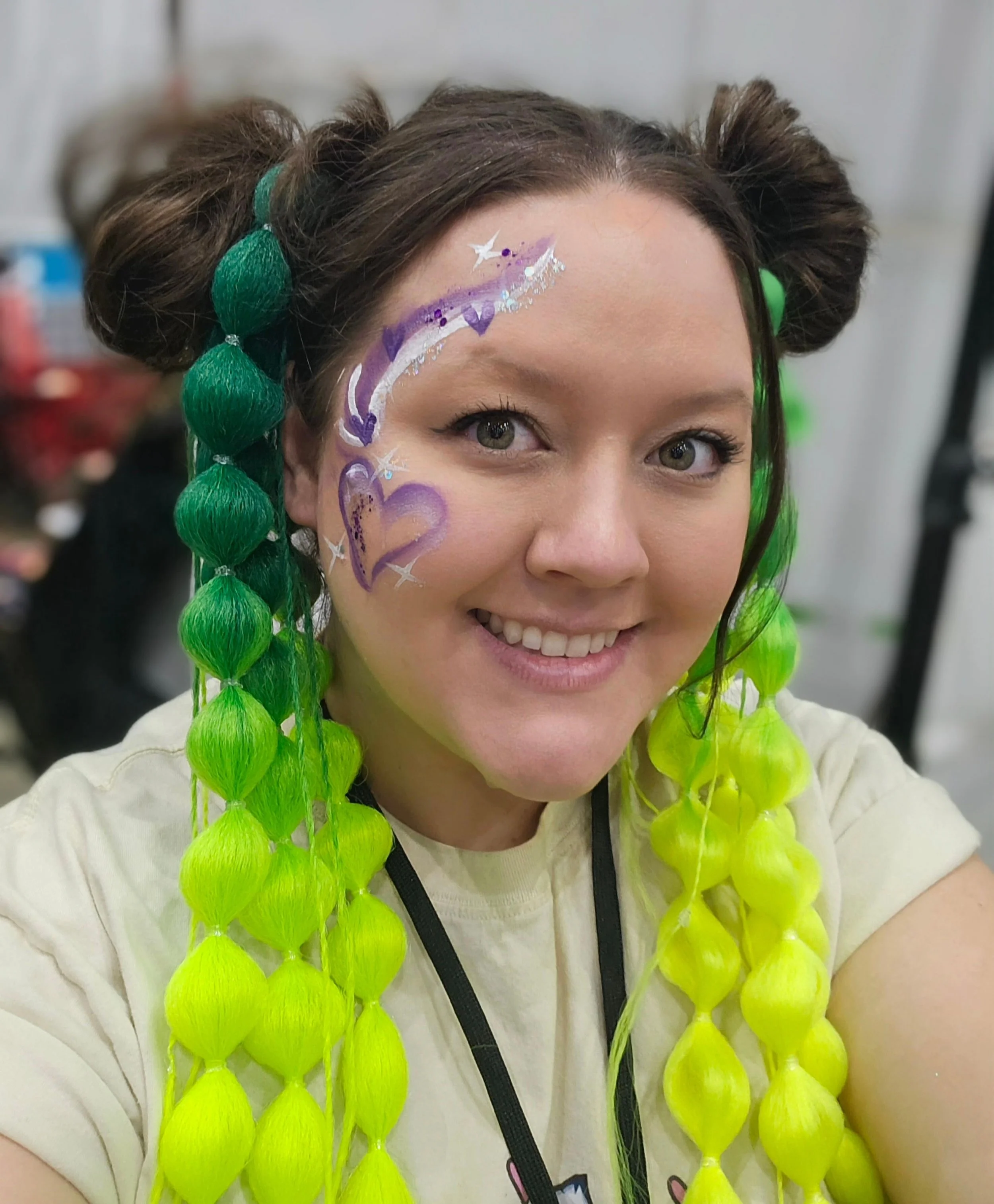 A woman with face paint of a purple and white unicorn and green hair styled in two buns. She is smiling and wearing a bright yellow and lime green lei.