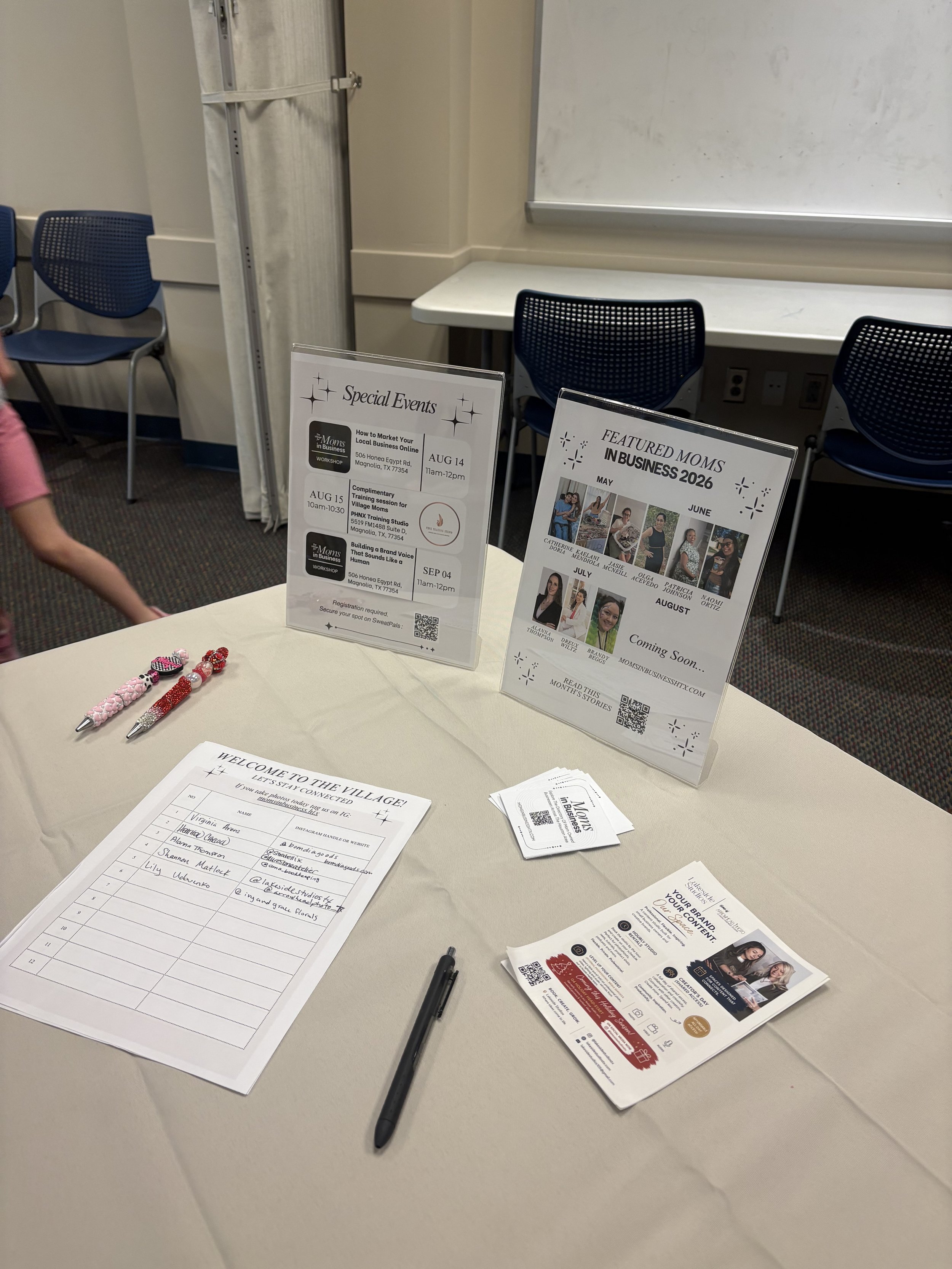 Table with pens, flyers, and signs about special events and business networking, in a conference room with chairs and whiteboard.