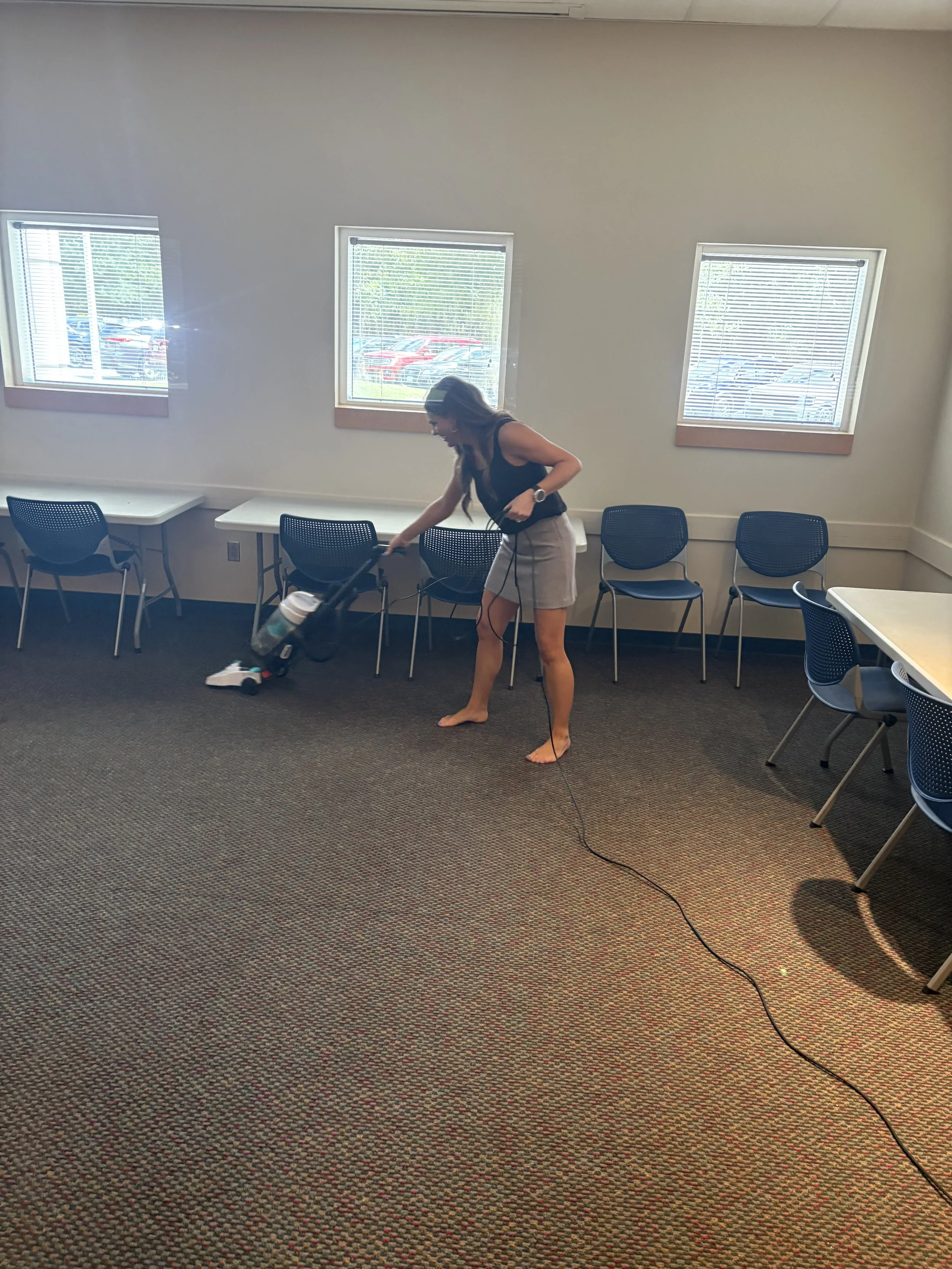 A woman with long hair, wearing a black top and beige shorts, polishing the floor with a mop in a room with gray carpet, white walls, and three windows with blinds, with chairs lined along the wall and a table in the corner.