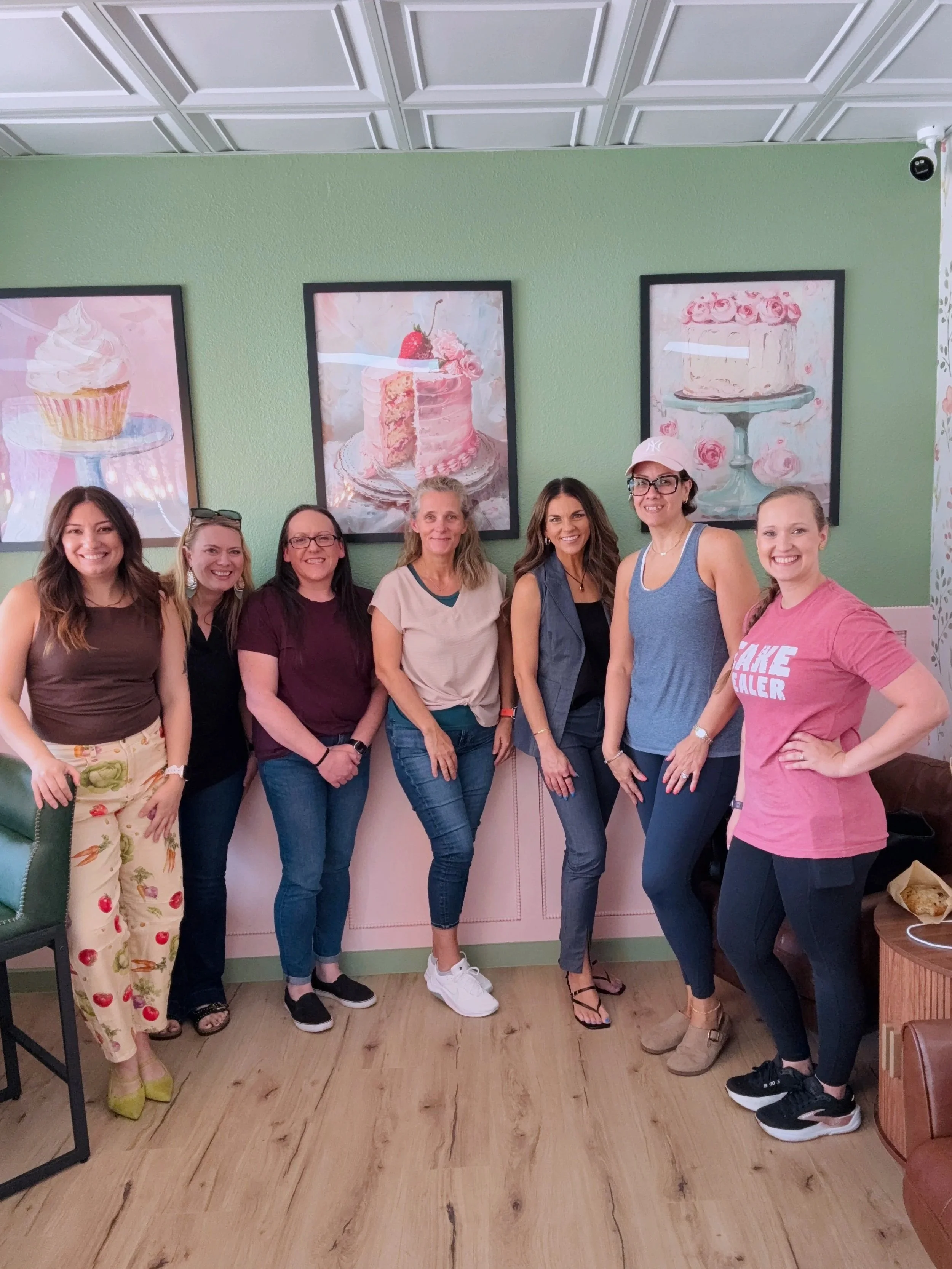 A group of eight women standing in a row inside a bakery or cafe, smiling at the camera. There are three framed pictures of cakes on a green wall behind them.