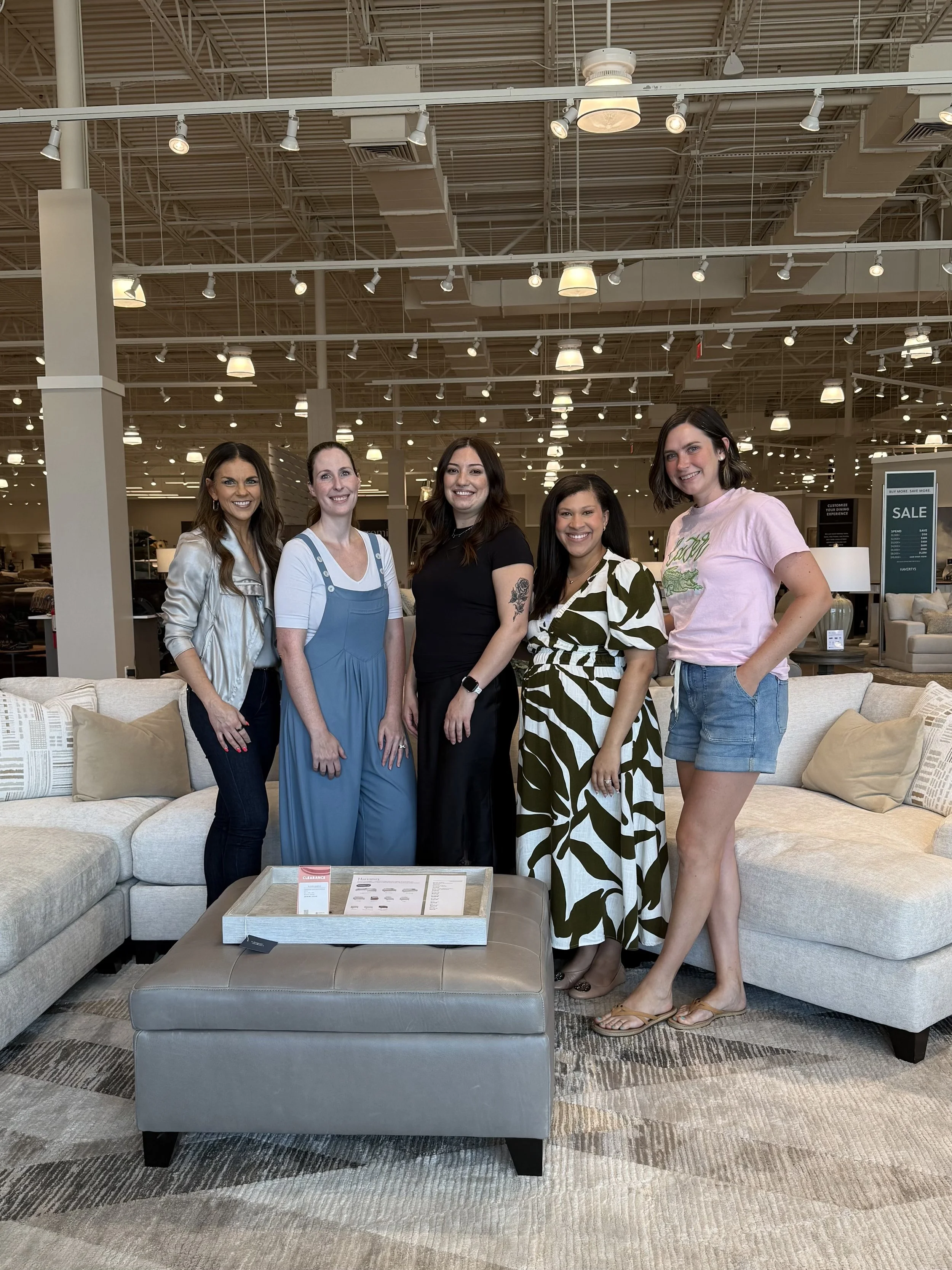 Five women standing in a furniture store, smiling for the camera, with a beige sofa and a tray table in front of them.