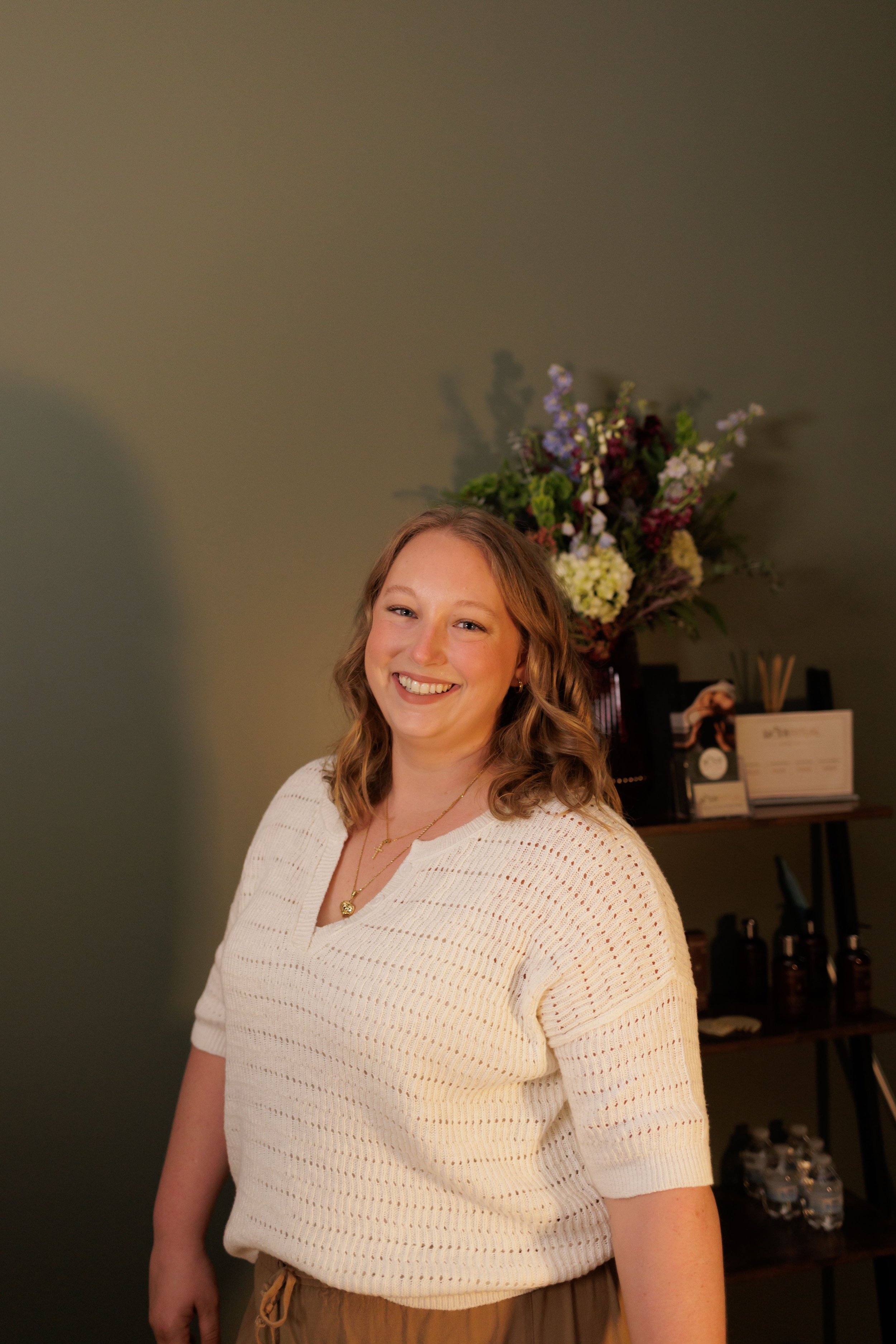 Smiling woman with light brown curly hair wearing a white knit sweater and gold jewelry, standing indoors with green wall and floral arrangement in the background.