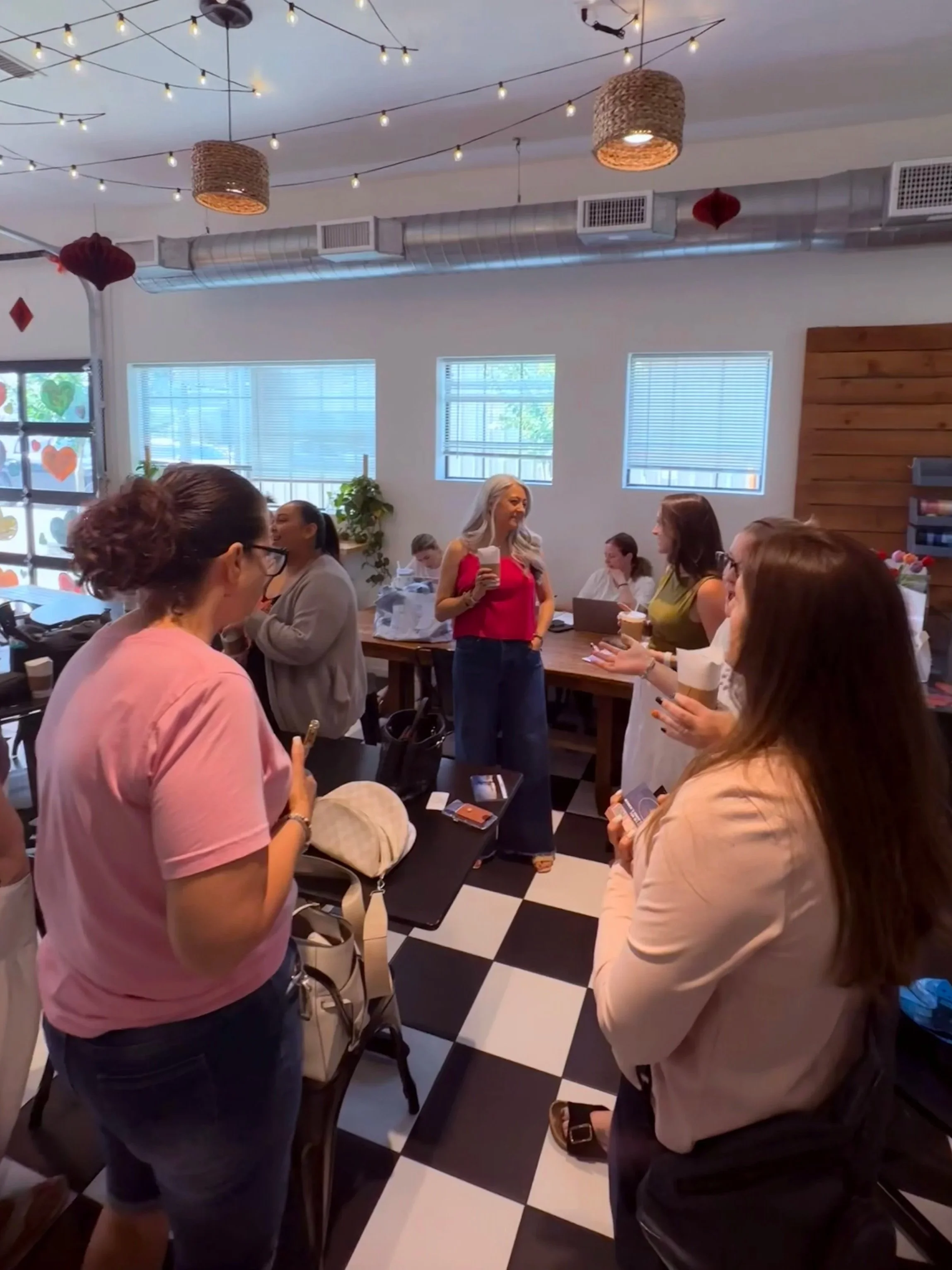 A group of women gathered inside a modern restaurant or cafe, engaging in conversation. The setting features white walls, multiple windows with blinds, string lights hanging from the ceiling, and a black and white checkered floor. Some women are holding drinks, and the atmosphere appears friendly and lively.