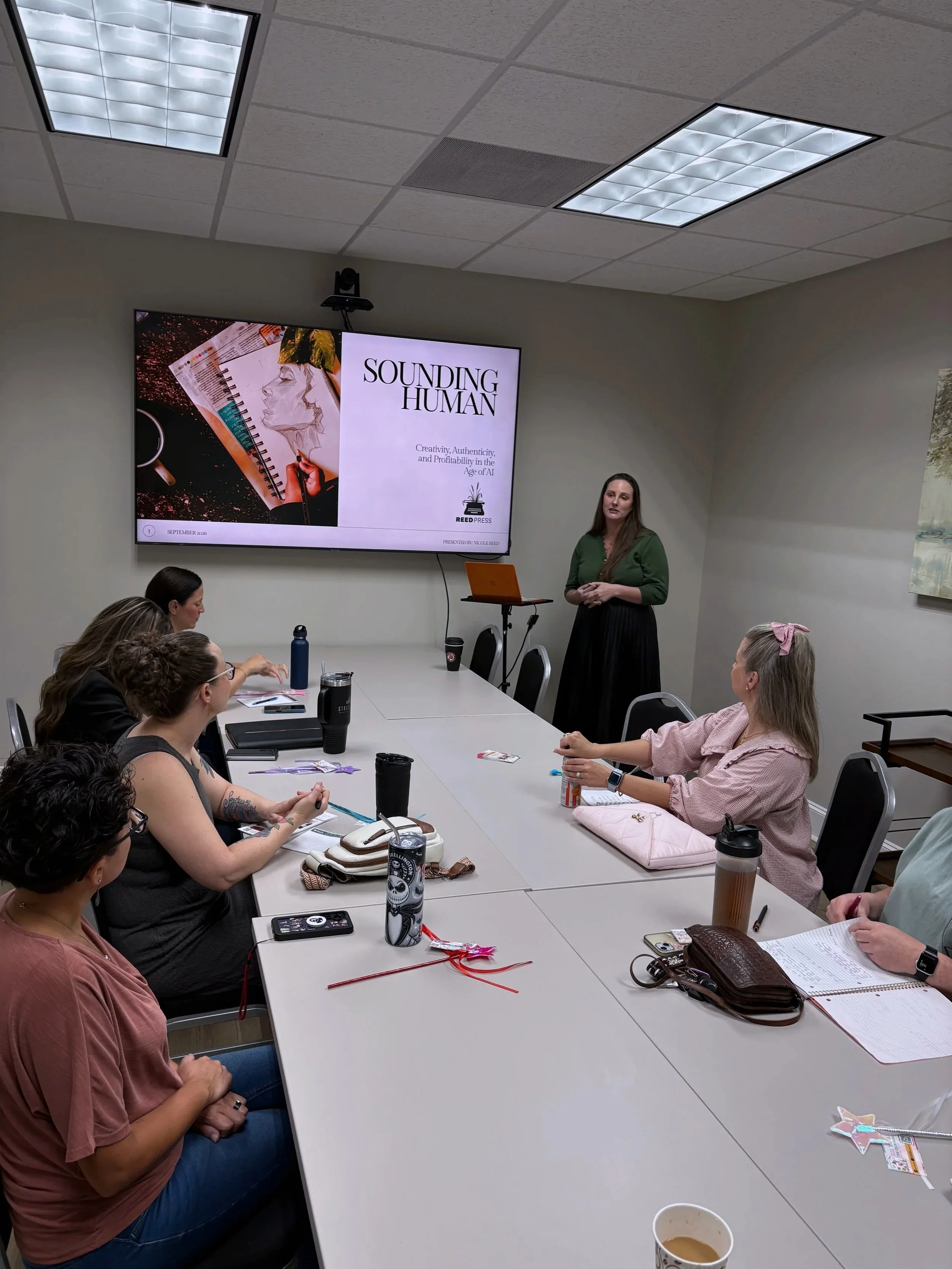 A woman is giving a presentation to a group of people in a conference room. The presentation slide reads 'Sounding Human' and discusses creativity, authenticity, and profitability in the age of AI. Several attendees are sitting at a long table with notebooks, drinks, and personal items, listening attentively.