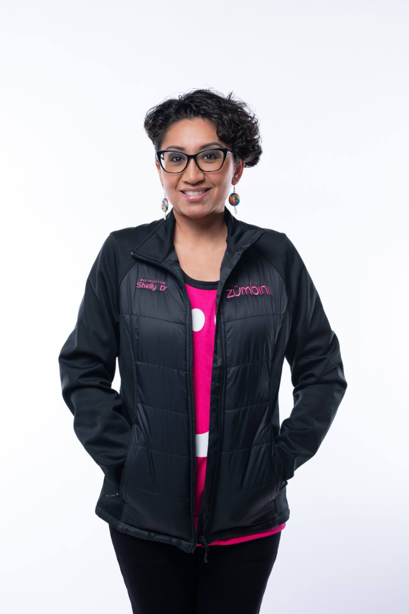 Woman with short curly dark hair, glasses, and colorful earrings, smiling, in a black jacket with pink accents, standing against a white background.