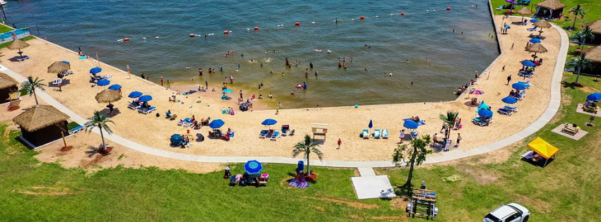 A crowded beach with people swimming and relaxing under blue umbrellas along a curved sandy shoreline. There are palm trees, a small pavilion, and grassy areas with more umbrellas and people nearby.