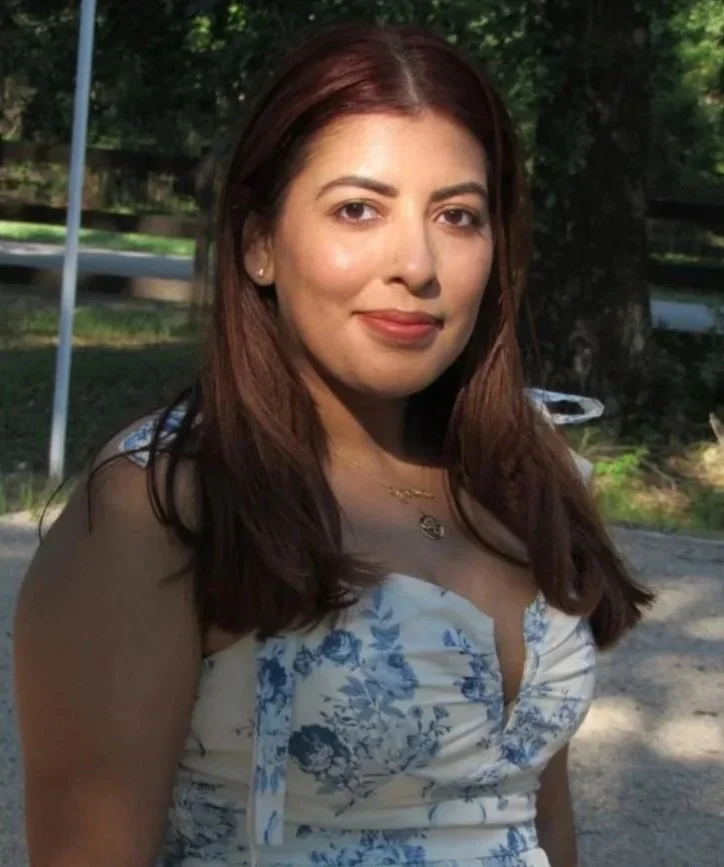 A woman with long brown hair wearing a white dress with blue floral prints, standing outdoors with trees and a fence in the background.