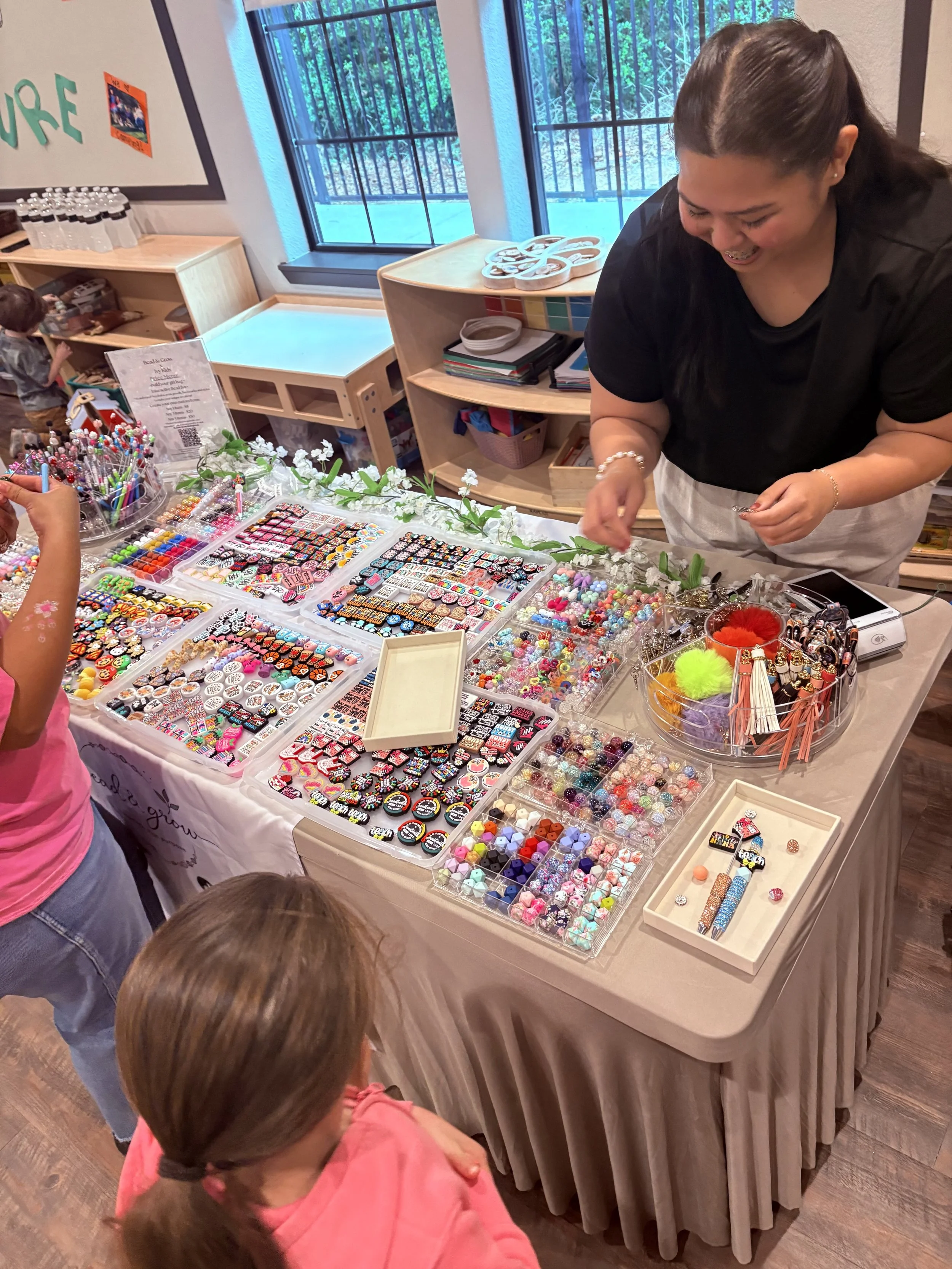 A young woman behind a table displaying colorful beads and jewelry making supplies at a craft fair.