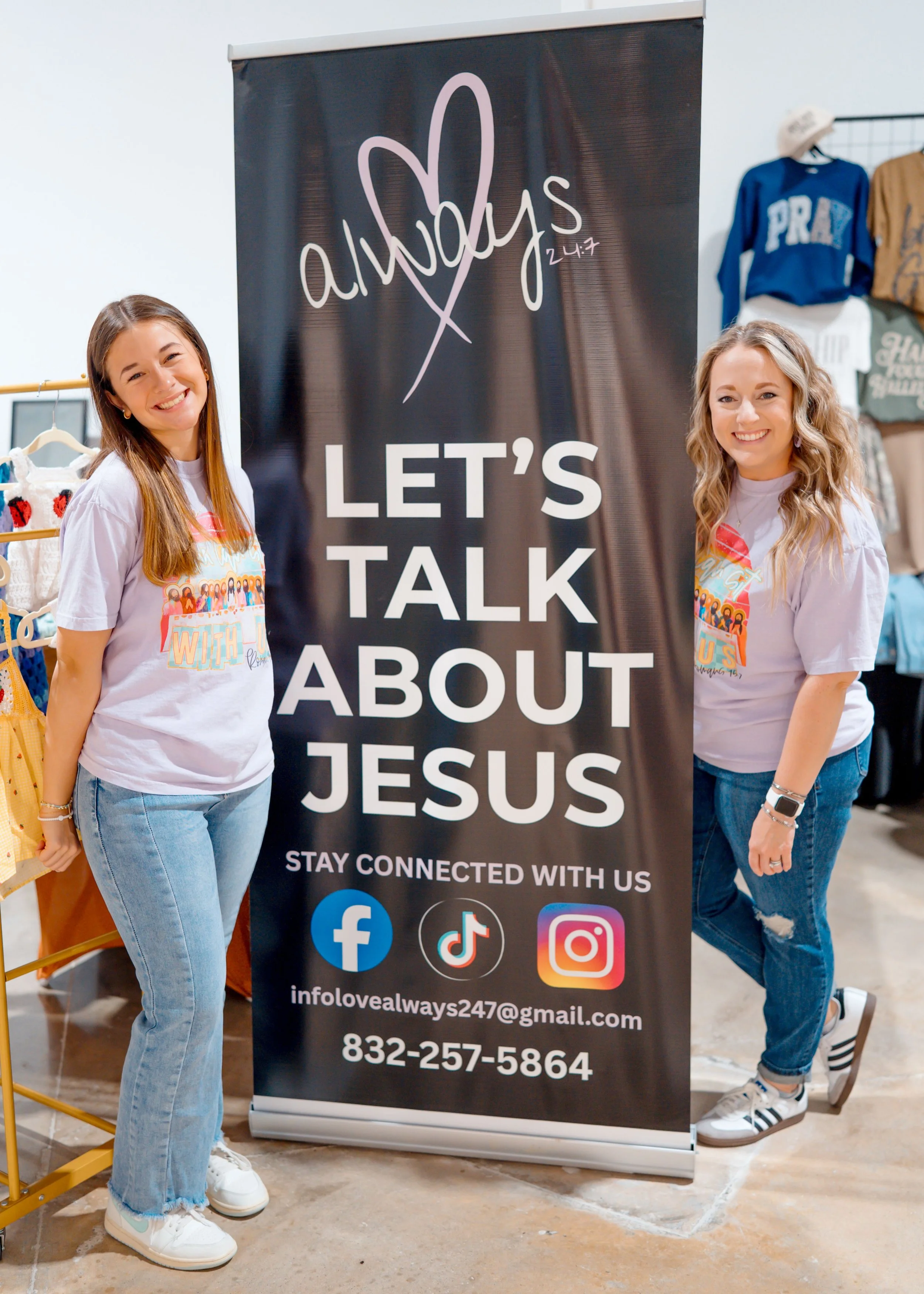 Two women standing inside a store next to a tall black banner with white and pink text promoting a social media outreach about Jesus, including icons for Facebook, TikTok, Instagram, and email, with a contact email and phone number at the bottom.
