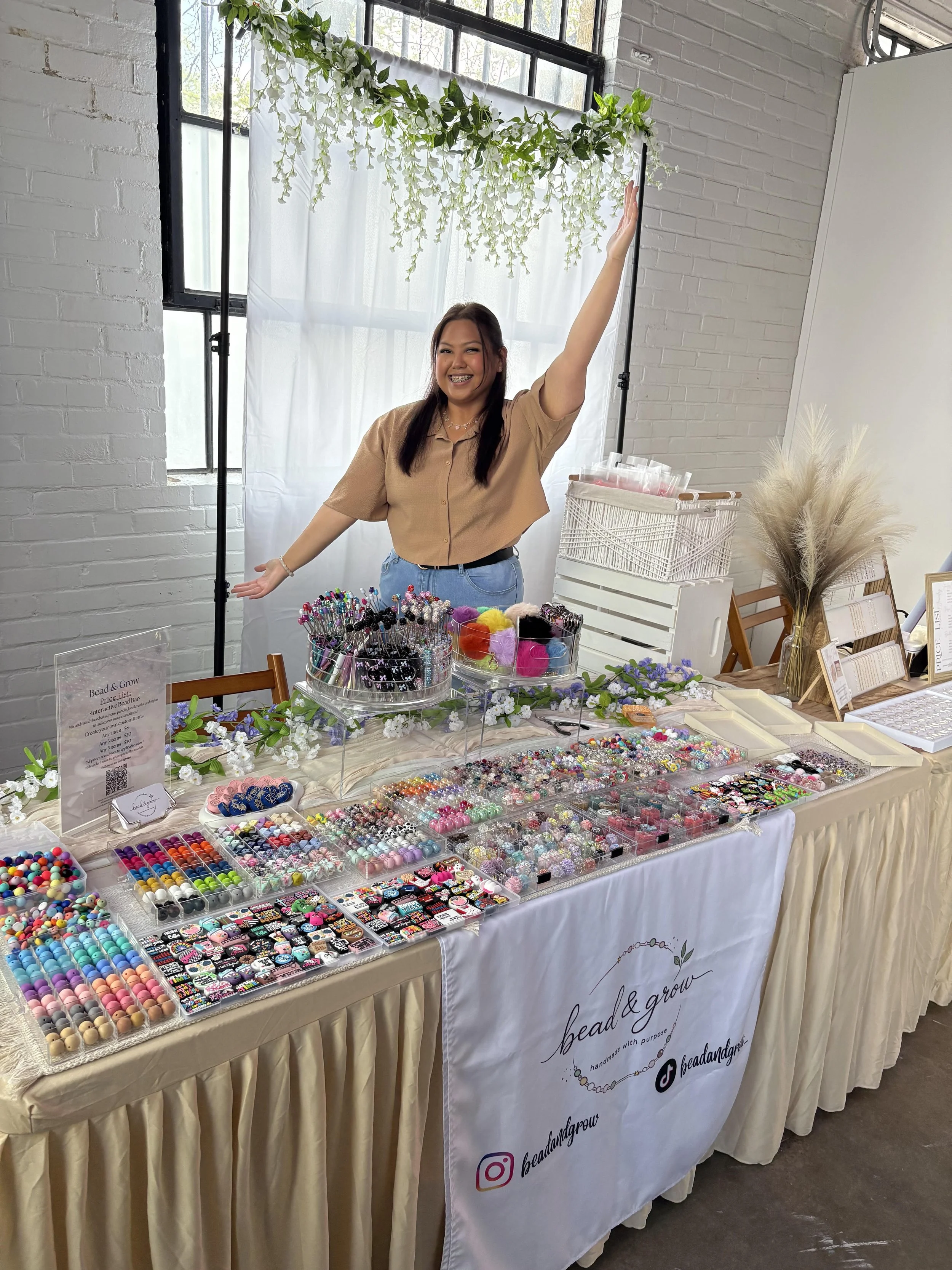 A smiling woman at a craft fair booth displaying a variety of colorful beads, buttons, and craft supplies, with a white brick wall, window, and decorative plants in the background.