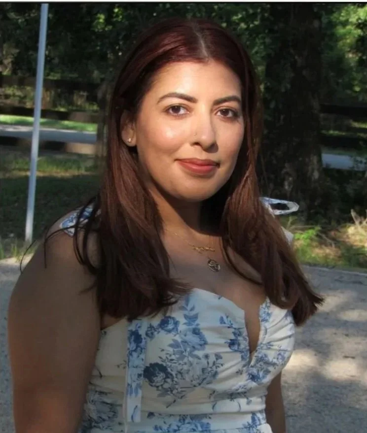 A woman with long brown hair, wearing a white dress with blue floral patterns, standing outdoors in a park with trees and a gravel path.