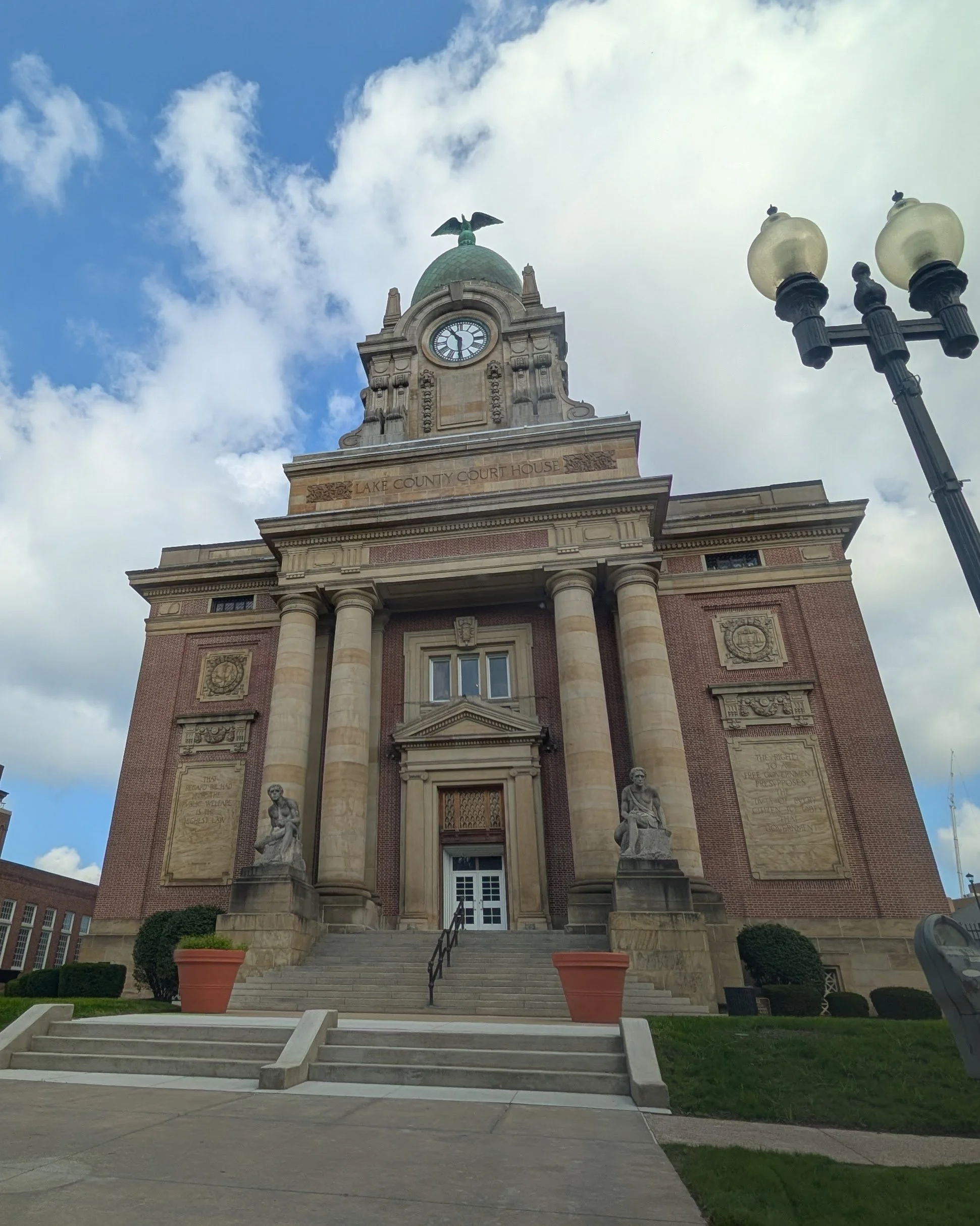 The historic Lake County courthouse with a clock tower topped by an eagle statue, featuring steps leading up to the entrance flanked by large potted plants, and two statues of seated figures at the base of the steps, under a partly cloudy sky.