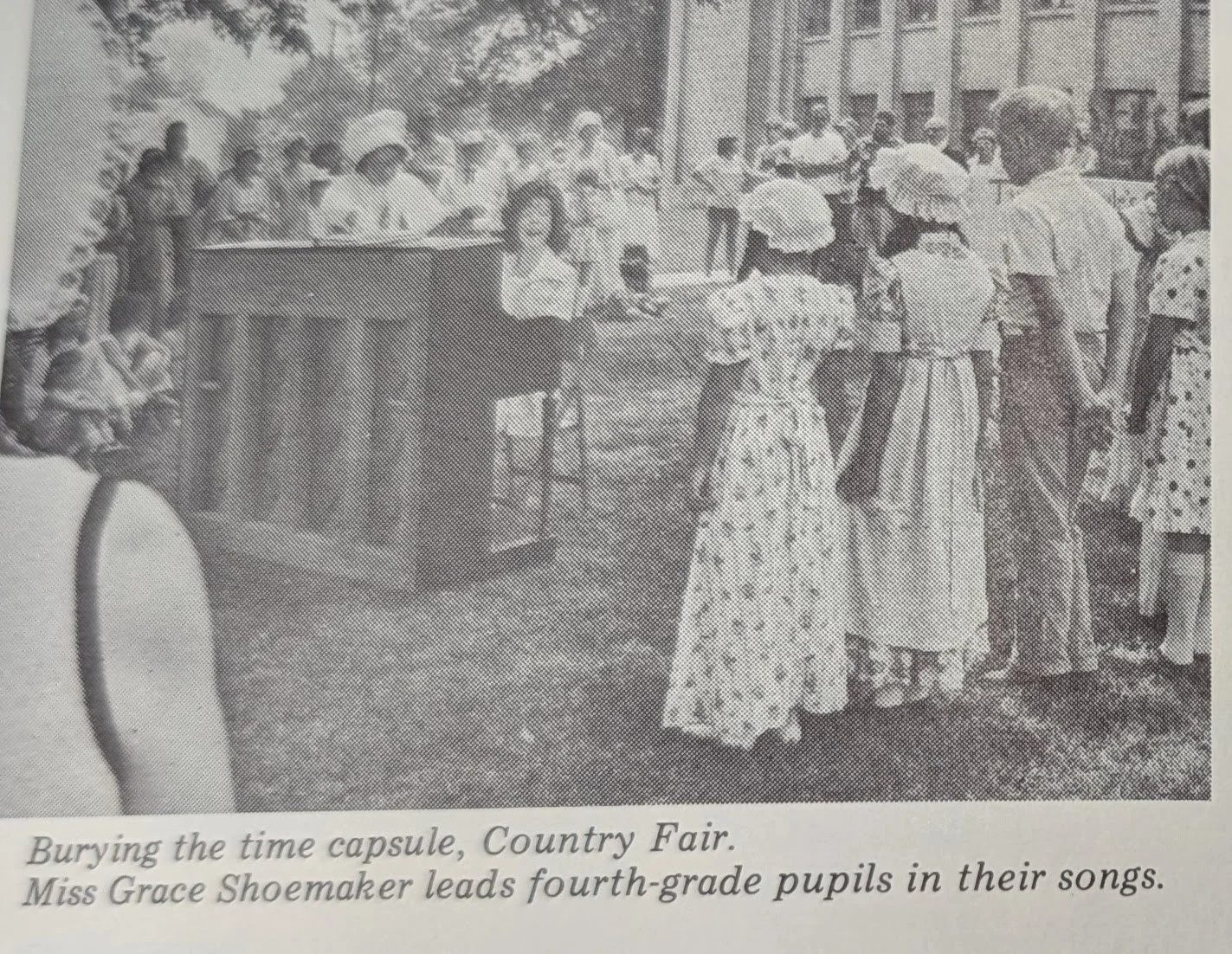 A black and white photo of children and women at a country fair, with some children singing and women wearing wide-brimmed hats standing in line. There are additional children on a float or platform decorated with streamers.