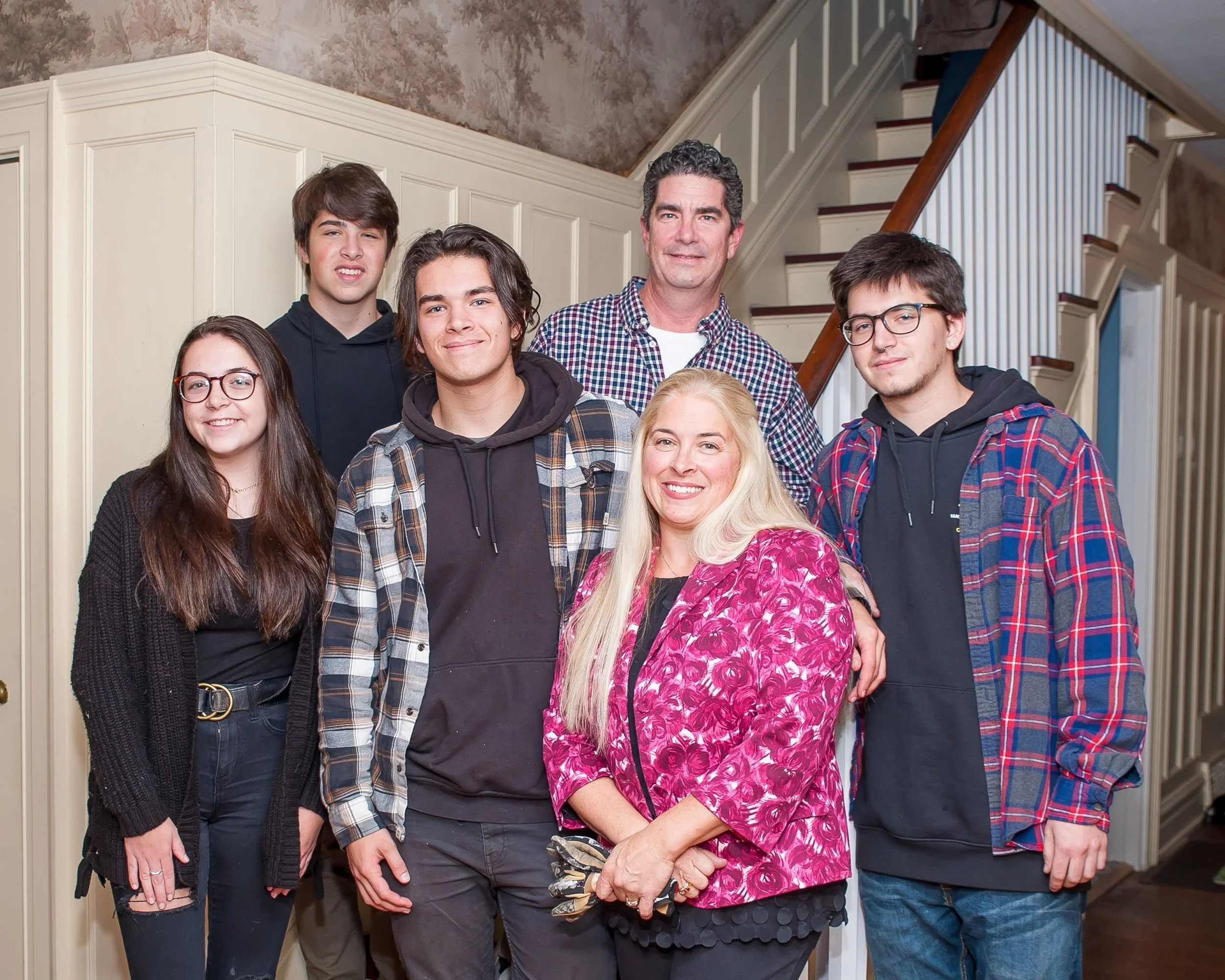 A group of six people, including teenagers and adults, standing together inside a house near a staircase with beige wainscoting and a textured wall. They are smiling and posing for the photo.
