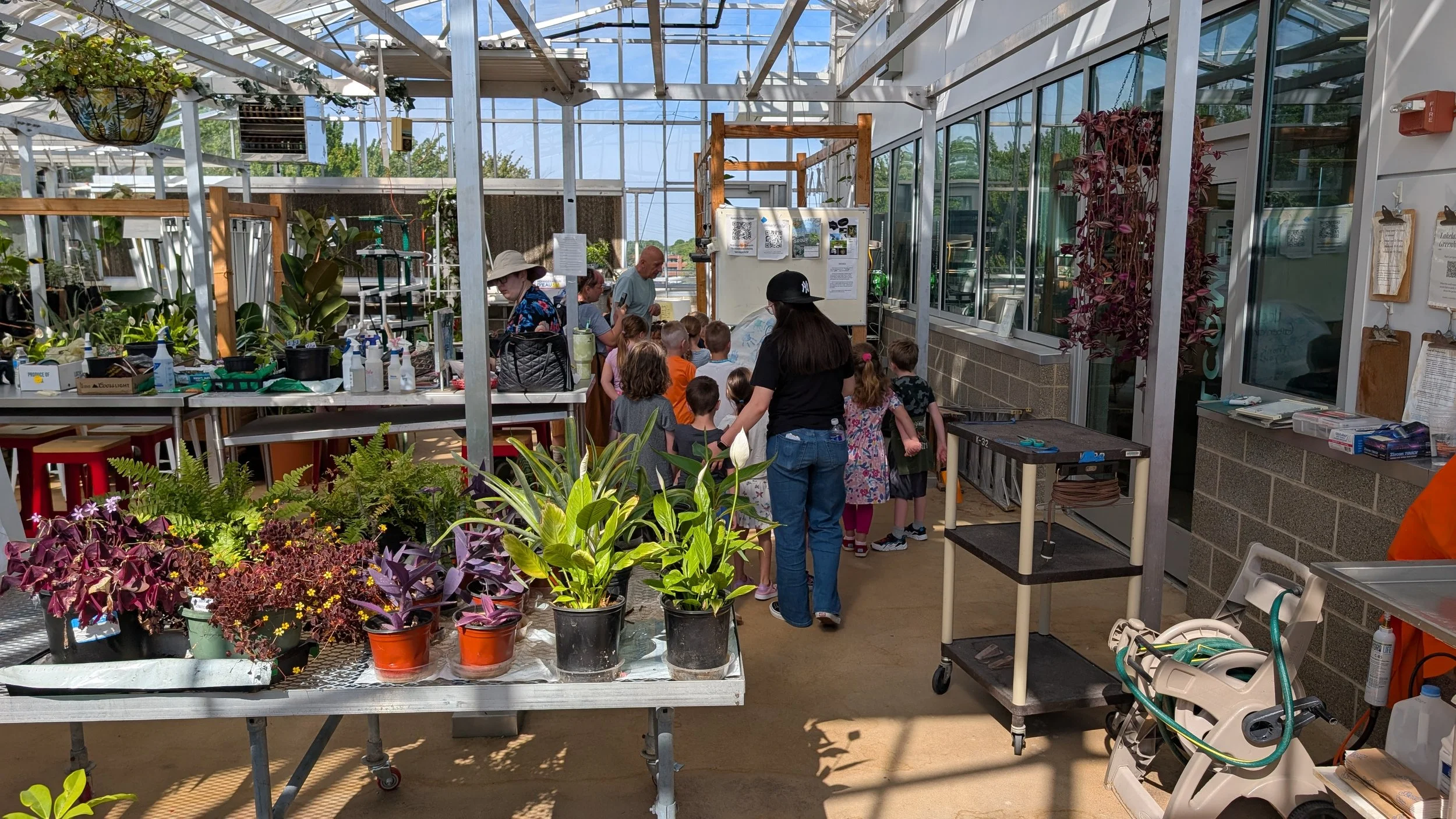 A group of children and adults in a greenhouse looking at plants and gardening supplies.