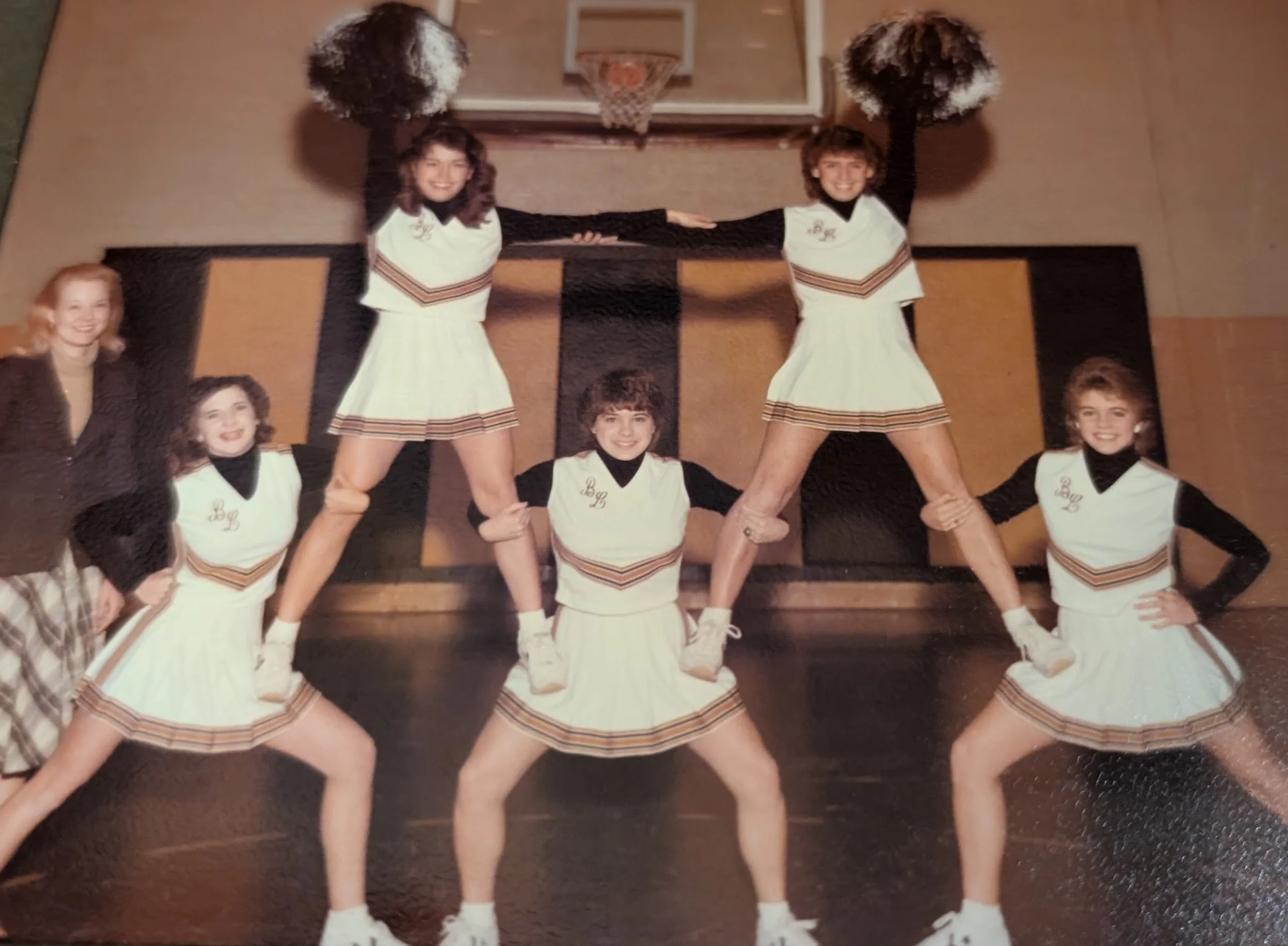 A vintage photo of five young women in cheerleader uniforms standing in a gymnasium with a basketball hoop in the background. They are arranged in a pyramid, smiling for the camera.