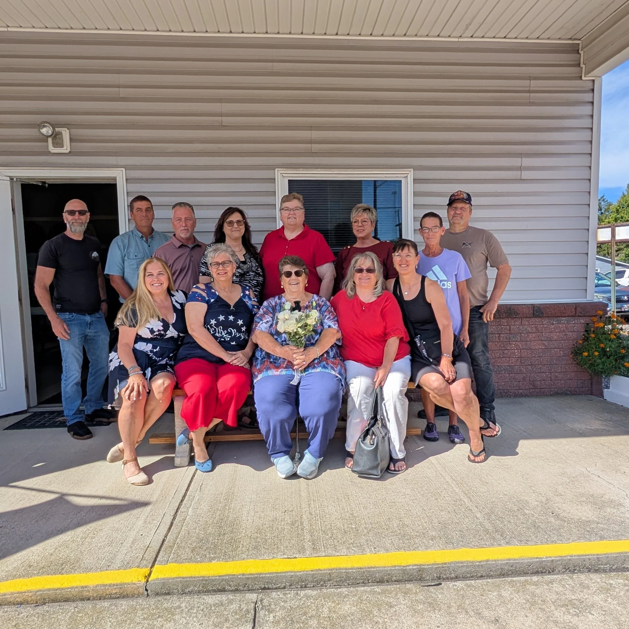 A group of people gathered for a photo outside a building, with some seated and others standing, in front of a beige building with a window and door, and a flower bed with orange flowers.