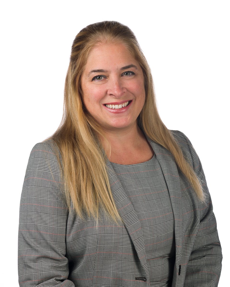 A woman with long blonde hair wearing a gray checkered blazer and dress, smiling at the camera.
