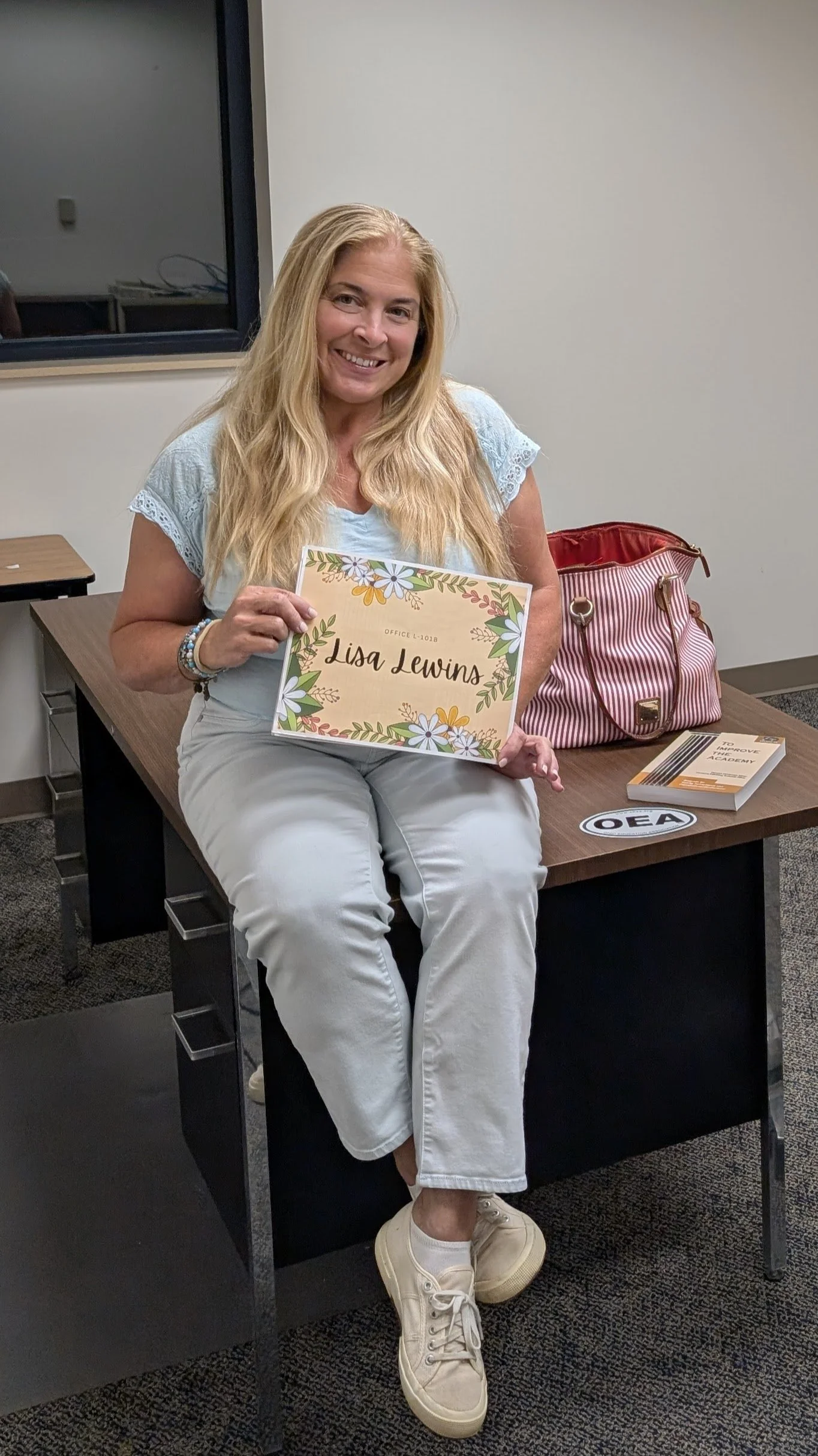 A woman sitting on a desk in an office, smiling, holding a nameplate that reads "Lisa Lewins" surrounded by floral designs. She has long blonde hair, wearing a light blue shirt with lace sleeves, white pants, and white sneakers. There is a red and white striped bag on the desk next to her, and a booklet titled "To Improve." There is a sticker with "OEA" on the desk. A window and a plain wall are in the background.