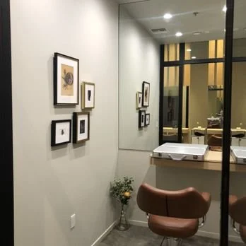 Interior of an art gallery or office area with framed pictures on the wall, a mirror, a small table with a flower arrangement, and a brown chair.