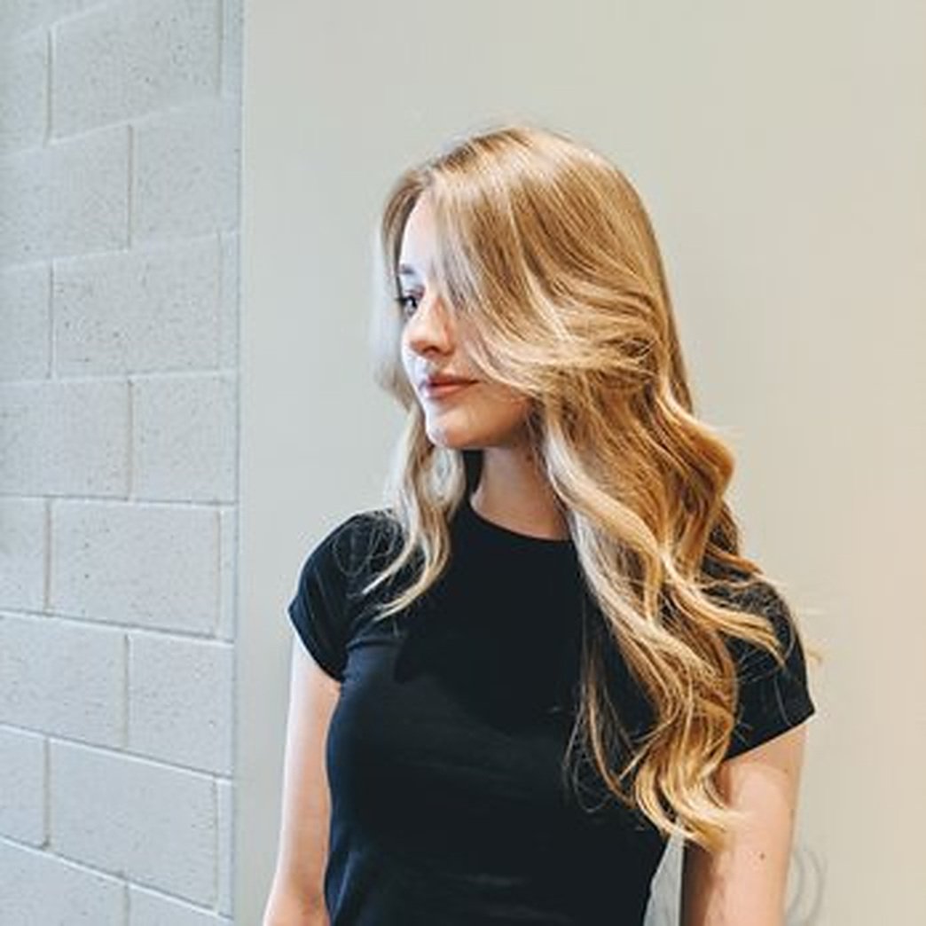 A young woman with long, wavy red hair wearing a black t-shirt standing against a wall.