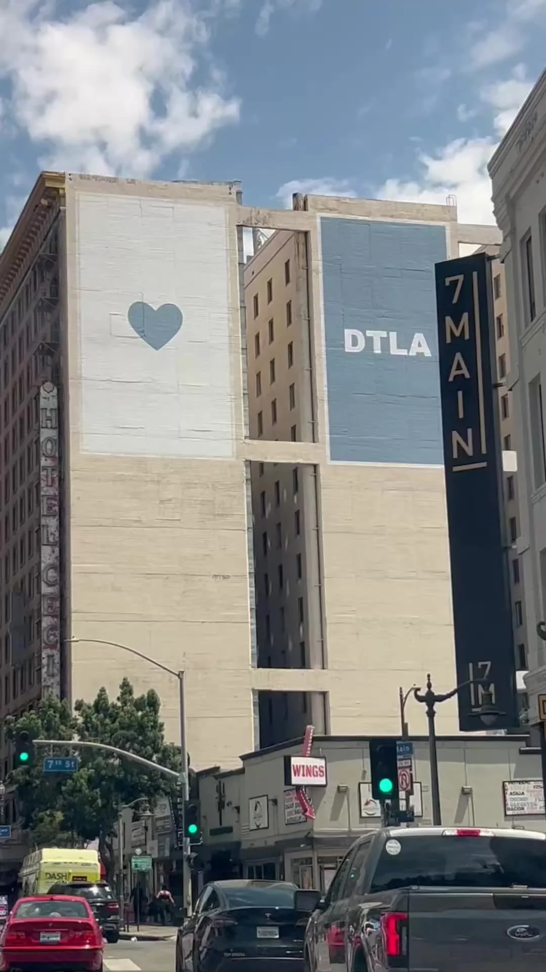 Buildings with murals, including a large white wall with a blue heart and the text 'DTLA' against a blue sky, on the corner of a city street with cars, traffic lights, and pedestrians.