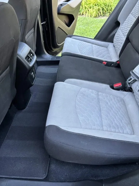 The backseat of a vehicle showing two seats with gray and black upholstery, seat belts, floor mats, and air vents near the center console. Outside greenery is visible through the open door.