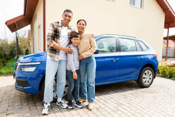 A happy family of three standing in front of their blue car parked on a driveway in front of a house. The family includes a man, a woman, and a young boy smiling at the camera.