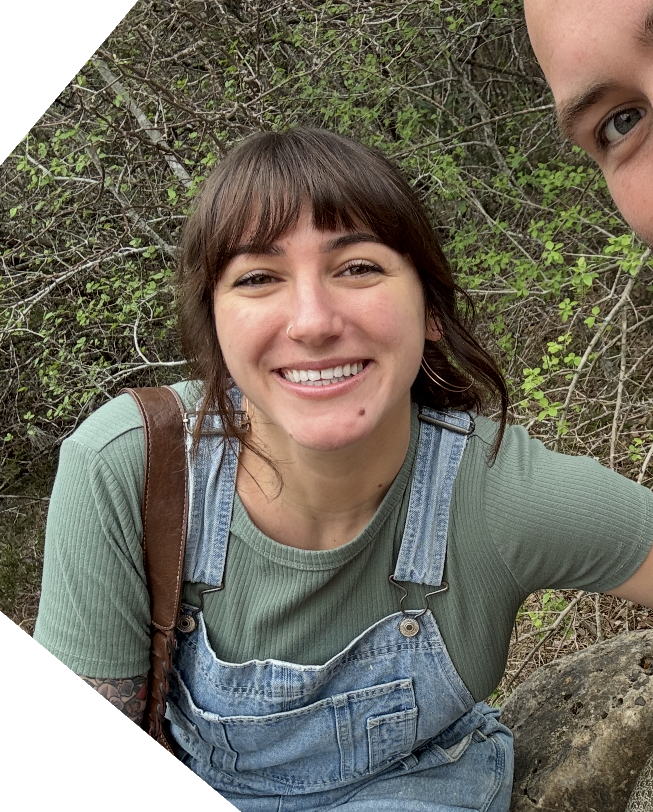 A woman with brown hair, wearing a green shirt, denim overalls, and hoop earrings, smiling outdoors in front of a background of green leaves and branches.