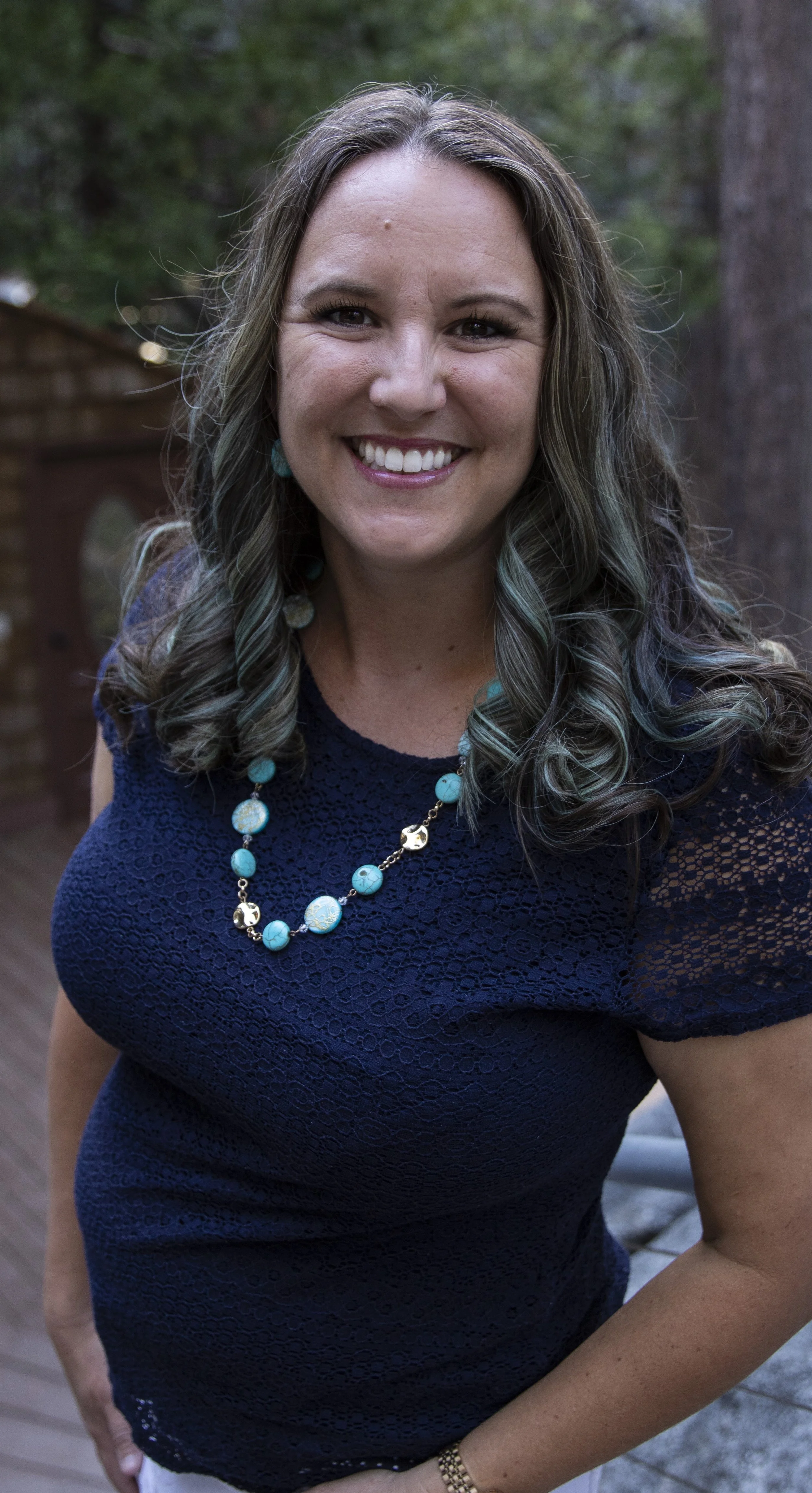 A woman with long, wavy, brown hair with highlights, wearing a navy blue lace dress and a turquoise bead necklace, standing outdoors next to trees.