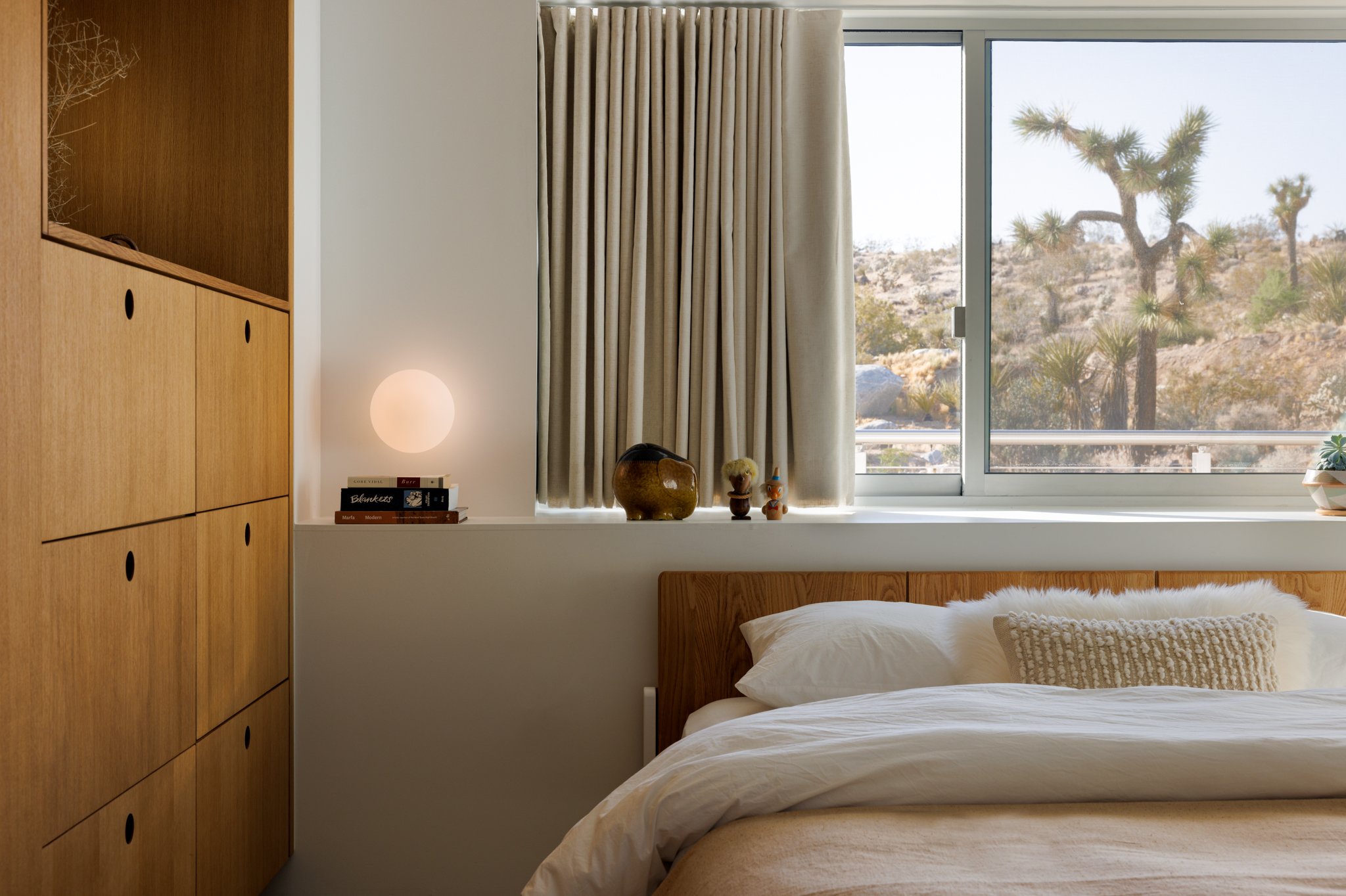 Bedroom at Panorama House featuring a custom solid white oak bed, Reform wardrobe cabinetry, Flos Glo-Ball wall light, and Fleetwood picture window framing a Joshua tree in the Mojave Desert.