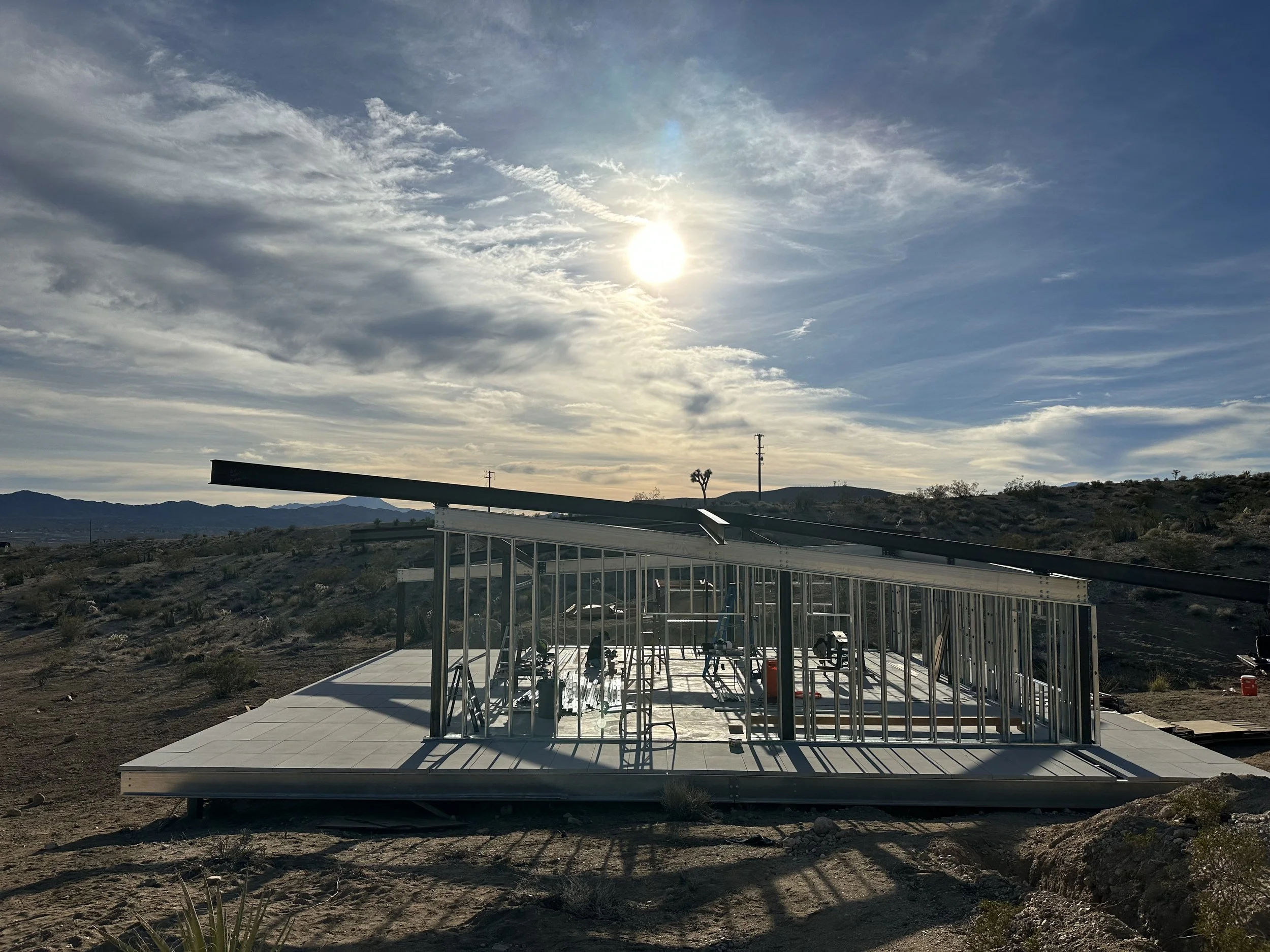 Panorama House under construction in Yucca Valley, showing its distinctive cantilevered roofline above exposed steel wall framing, with the Mojave Desert mountains and afternoon sun beyond.
