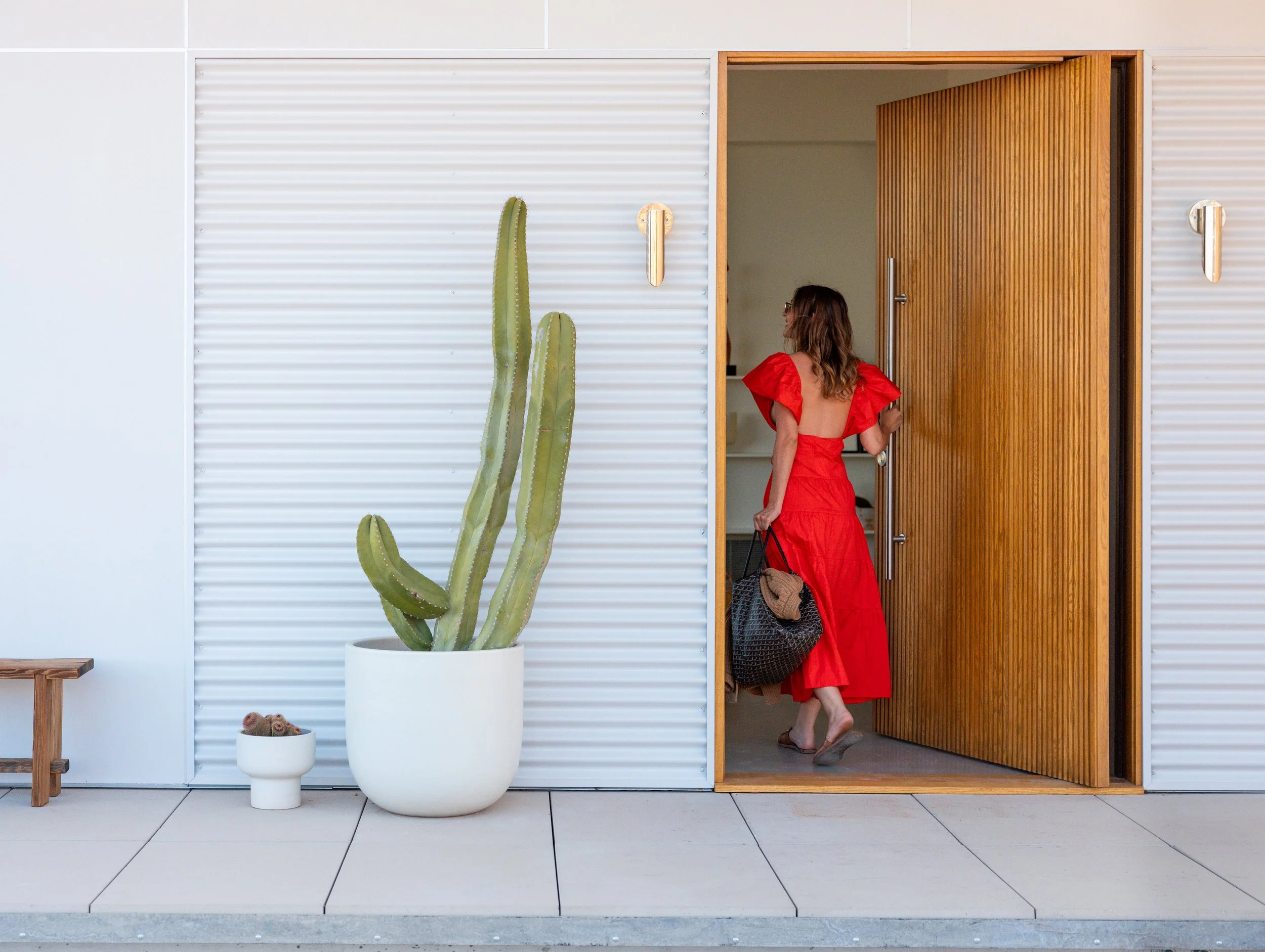 Woman in a red dress entering Panorama House through a custom white oak pivot door, beside a tall cactus in a Case Study planter and Everett LED wall sconces on the white exterior.