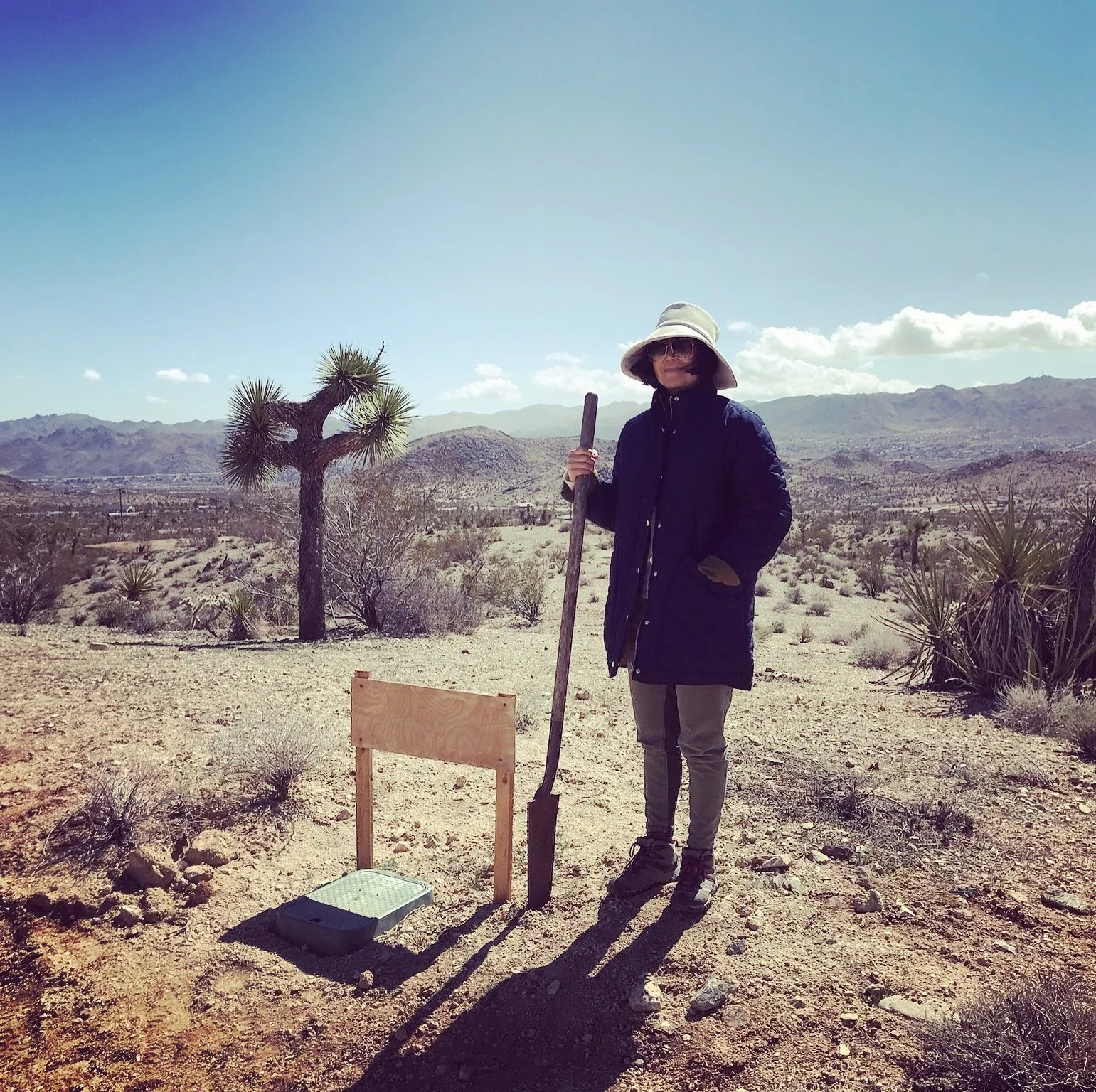 Panorama House owner standing with a shovel on the undeveloped desert property in Yucca Valley, surrounded by Joshua trees and distant mountains.