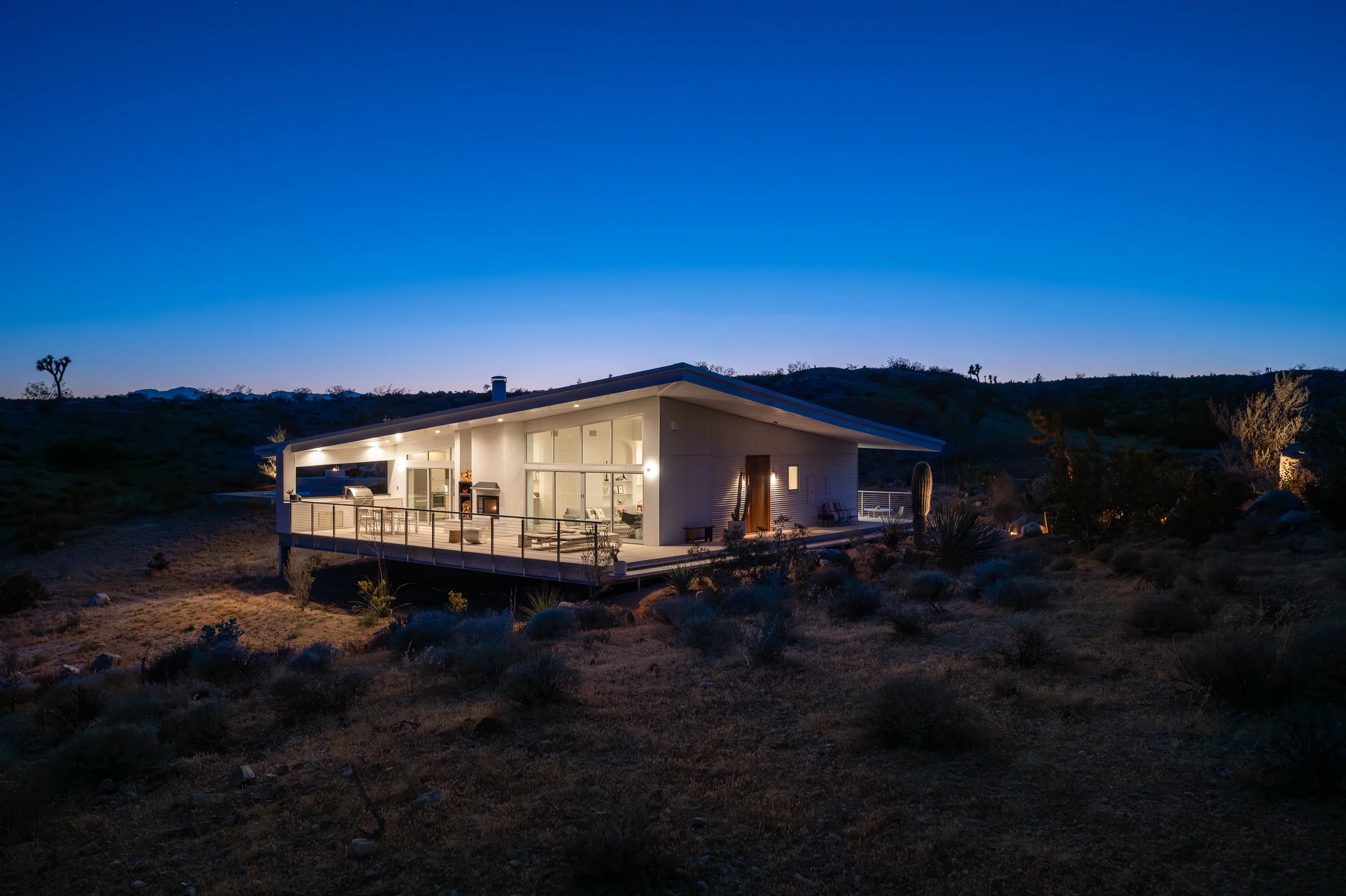 Panorama House illuminated at blue hour in Yucca Valley, with expansive Fleetwood sliding glass doors, a wraparound deck, and its distinctive folding roofline against the Mojave Desert landscape.