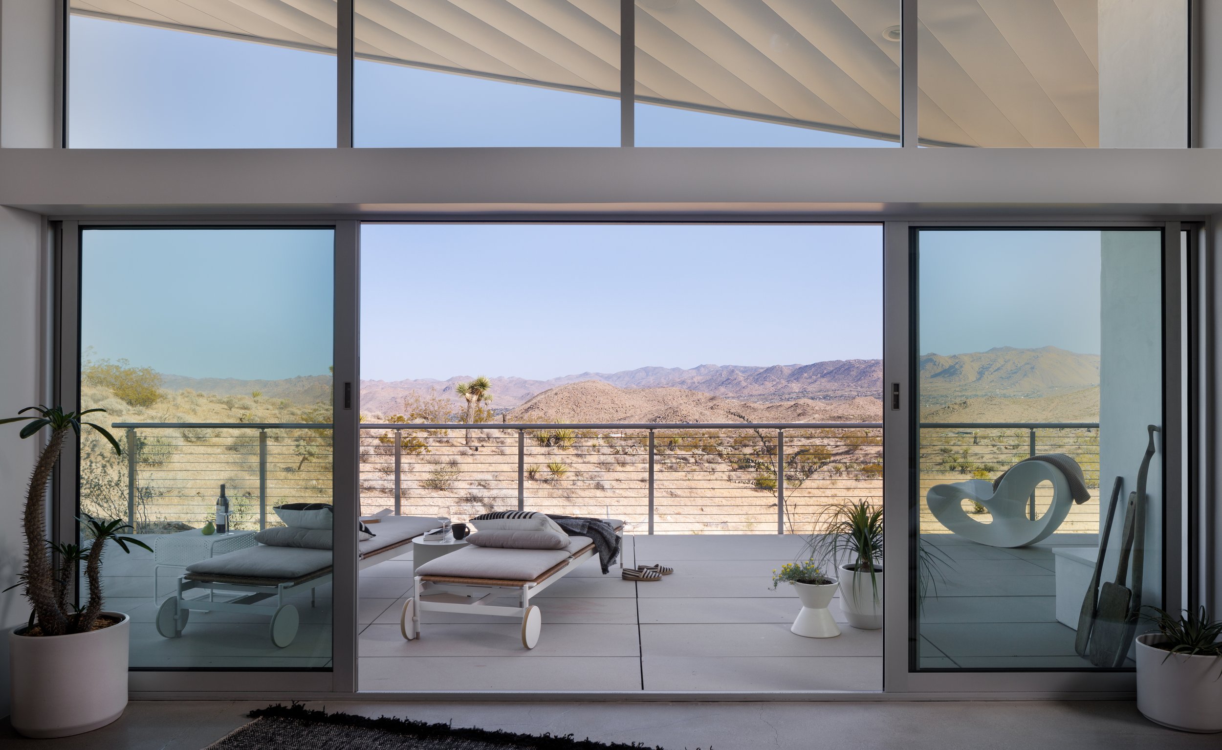 View from the living room of Panorama House through floor-to-ceiling Fleetwood sliding glass doors onto the wraparound deck, with outdoor lounge chairs and panoramic Mojave Desert views.