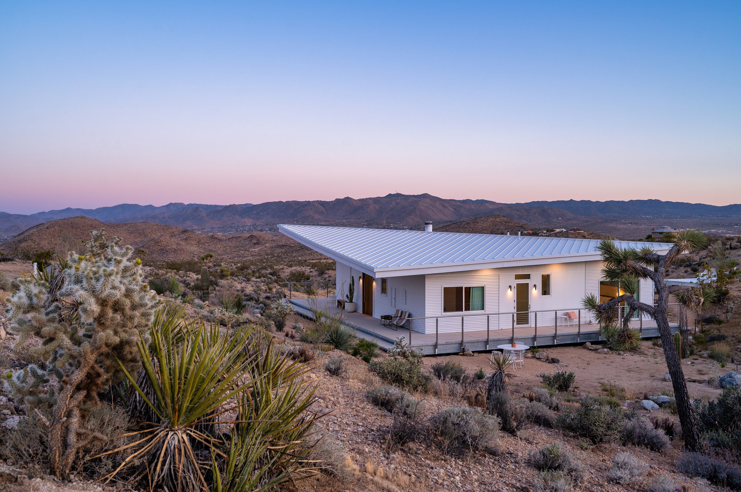 Panorama House at dusk in Yucca Valley, California, with its distinctive folding standing-seam metal roof and wraparound deck, surrounded by Joshua trees and Mojave Desert mountains.