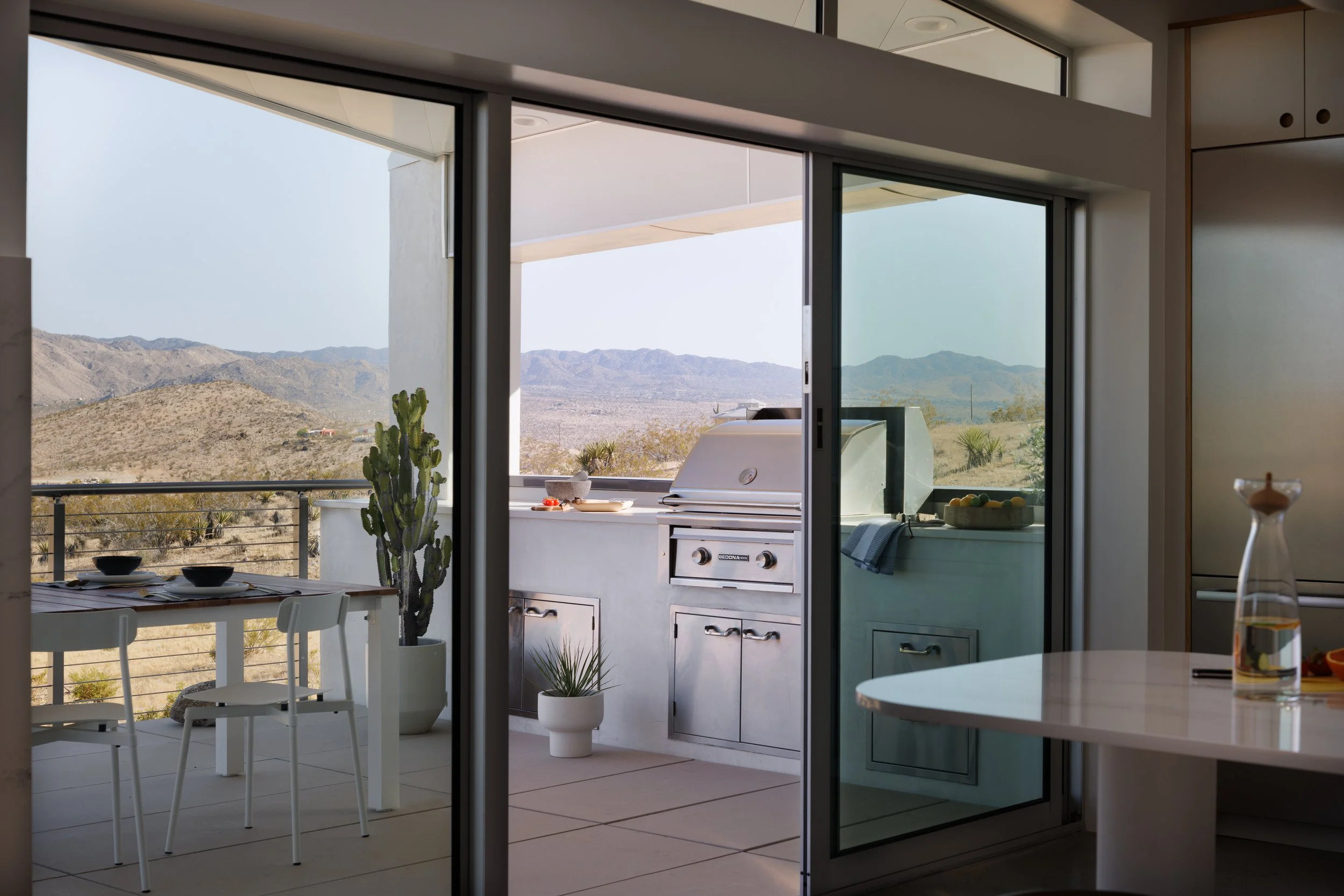 View from the Panorama House kitchen through Fleetwood sliding glass doors onto the deck, featuring a Lynx outdoor grill, Oasiq teak dining table, Petite Friture Fromme chairs, and panoramic Mojave Desert mountain views.