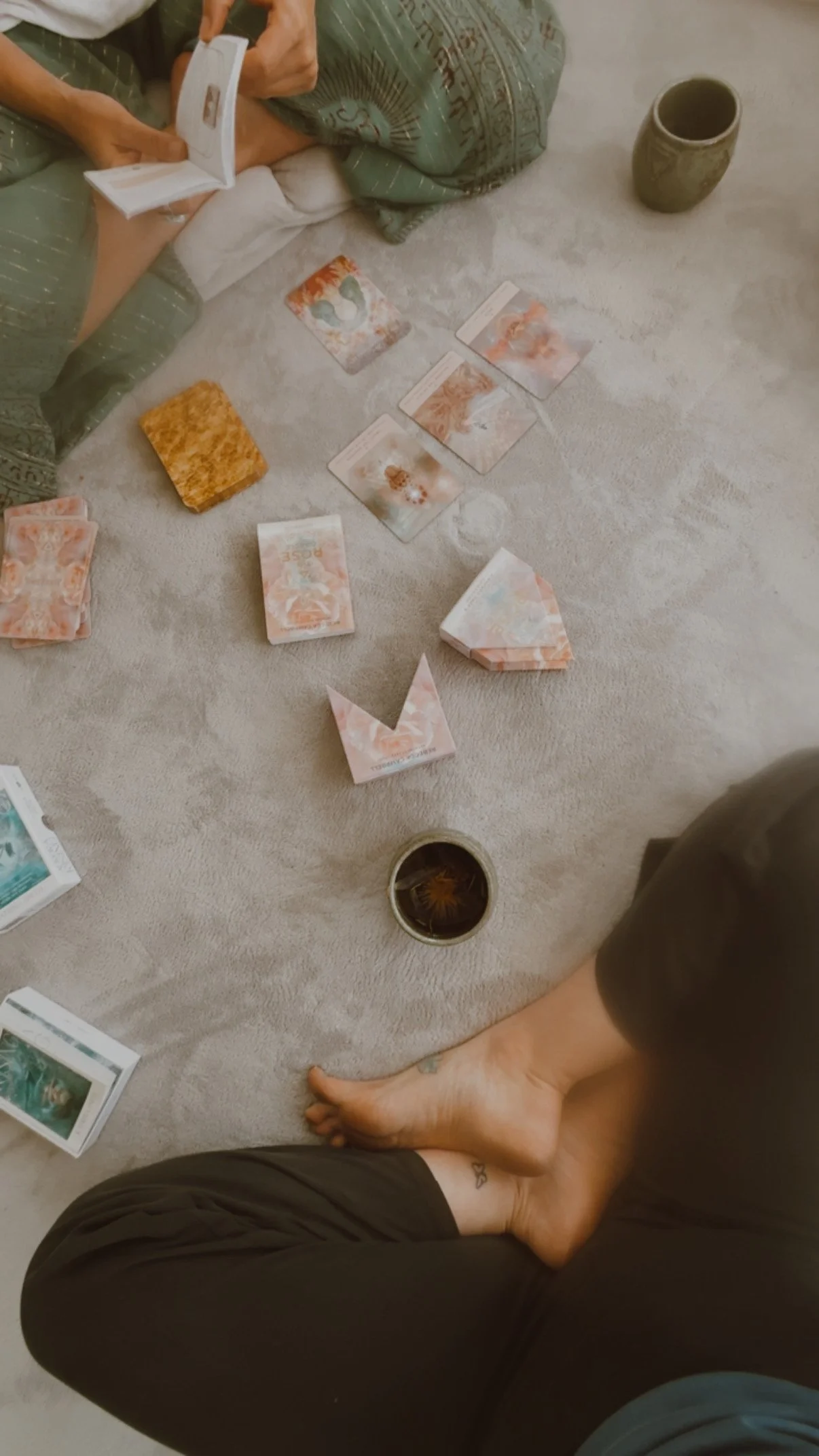 A person sitting on the floor with legs crossed, reading a tarot card deck, with several tarot cards laid out on a light-colored carpet. There are also cups and additional tarot cards nearby.
