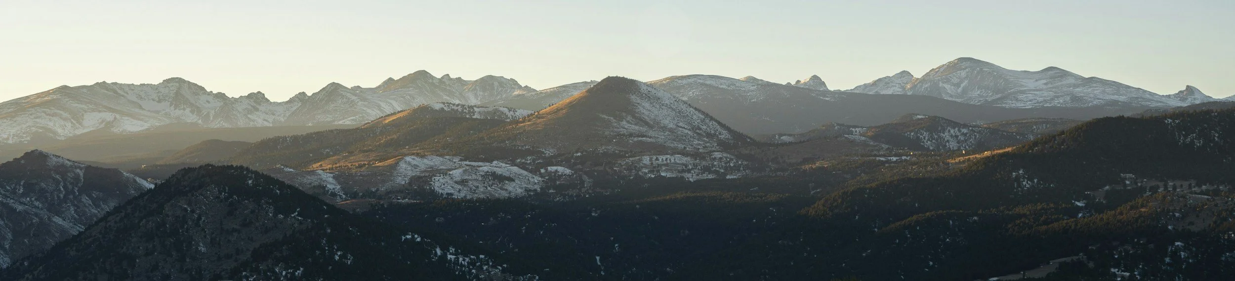 Mountain range with snow-capped peaks and green hills under a clear sky during sunset.
