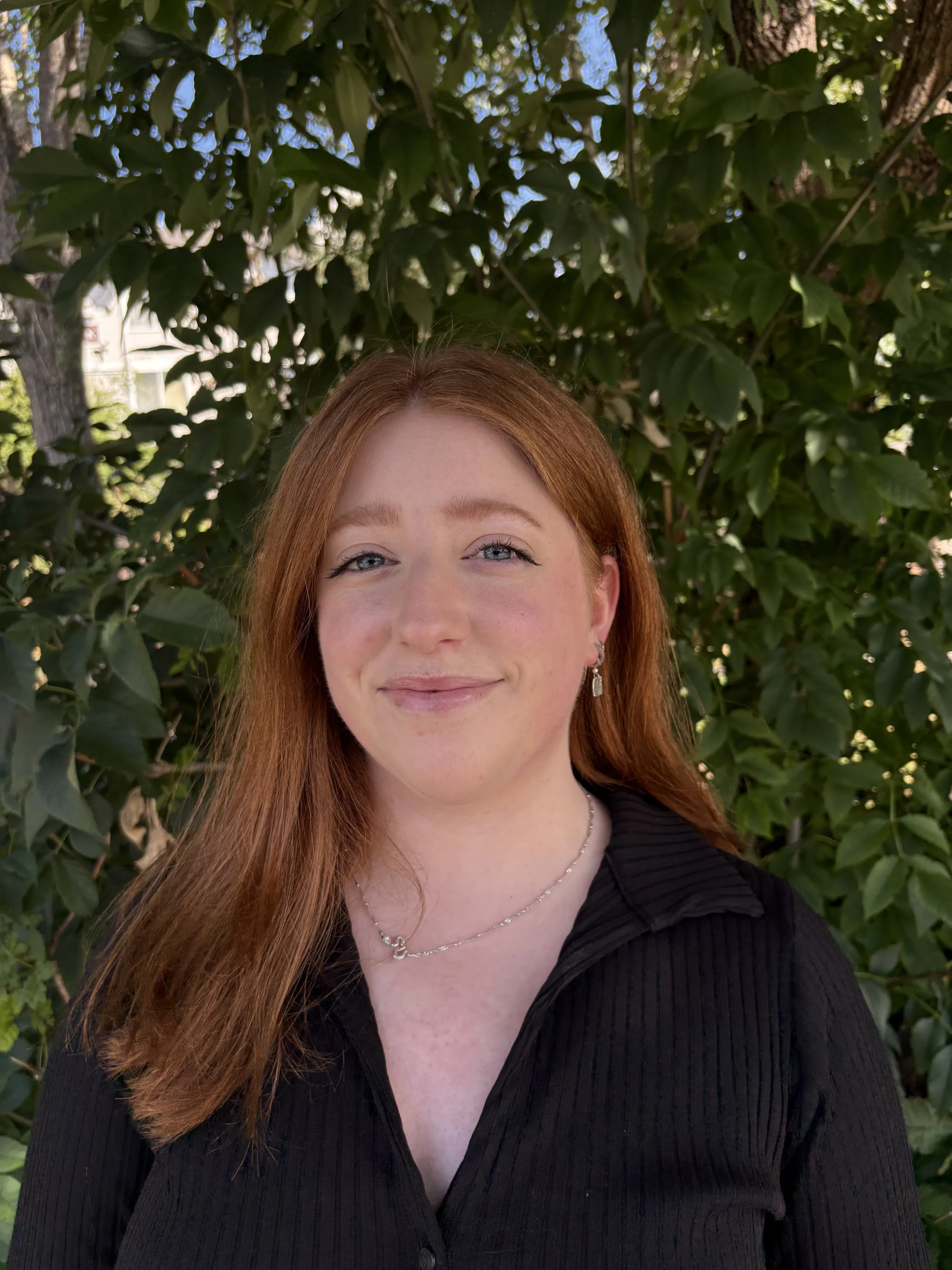 A woman with long red hair and light skin smiling outdoors in front of green foliage, wearing a black ribbed shirt, earrings, and a necklace.