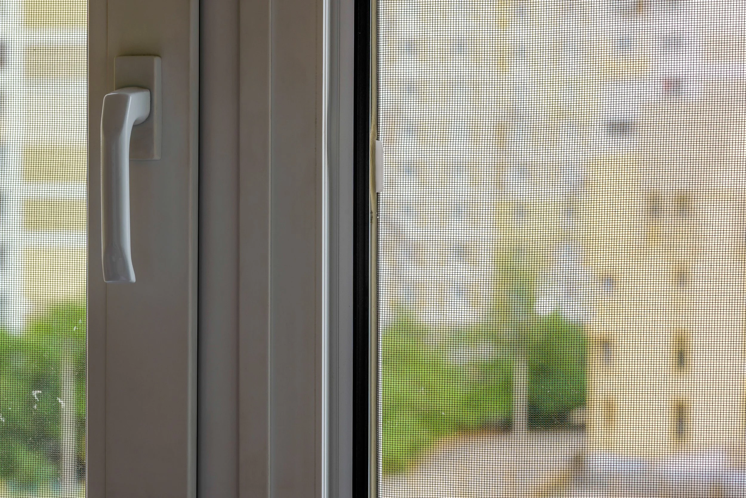 Close-up of a white window screen with a white window handle and a view of a green outdoor space.
