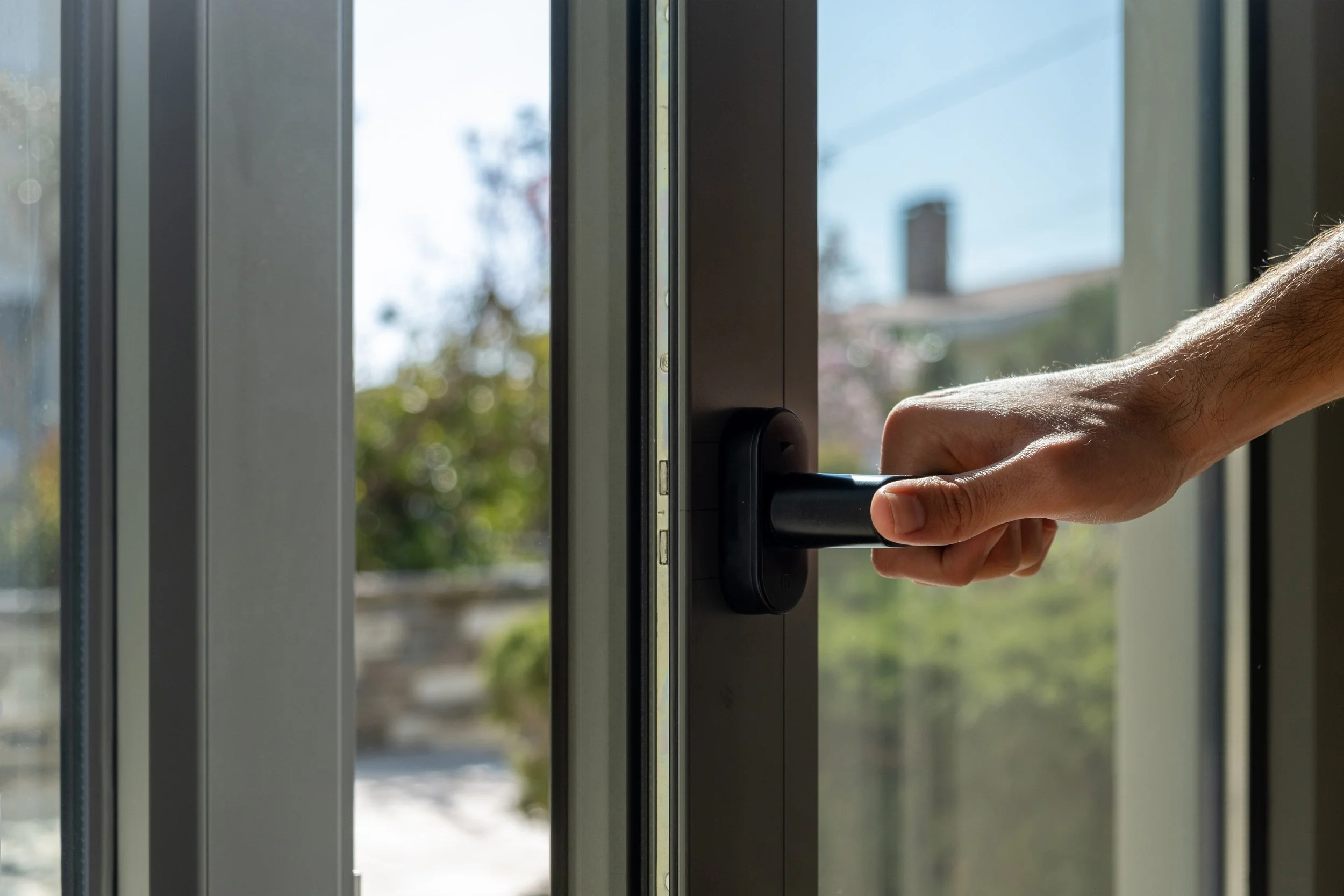 A person's hand opening a glass door with a black handle, sunlight outside, trees and a house chimney visible in the background.